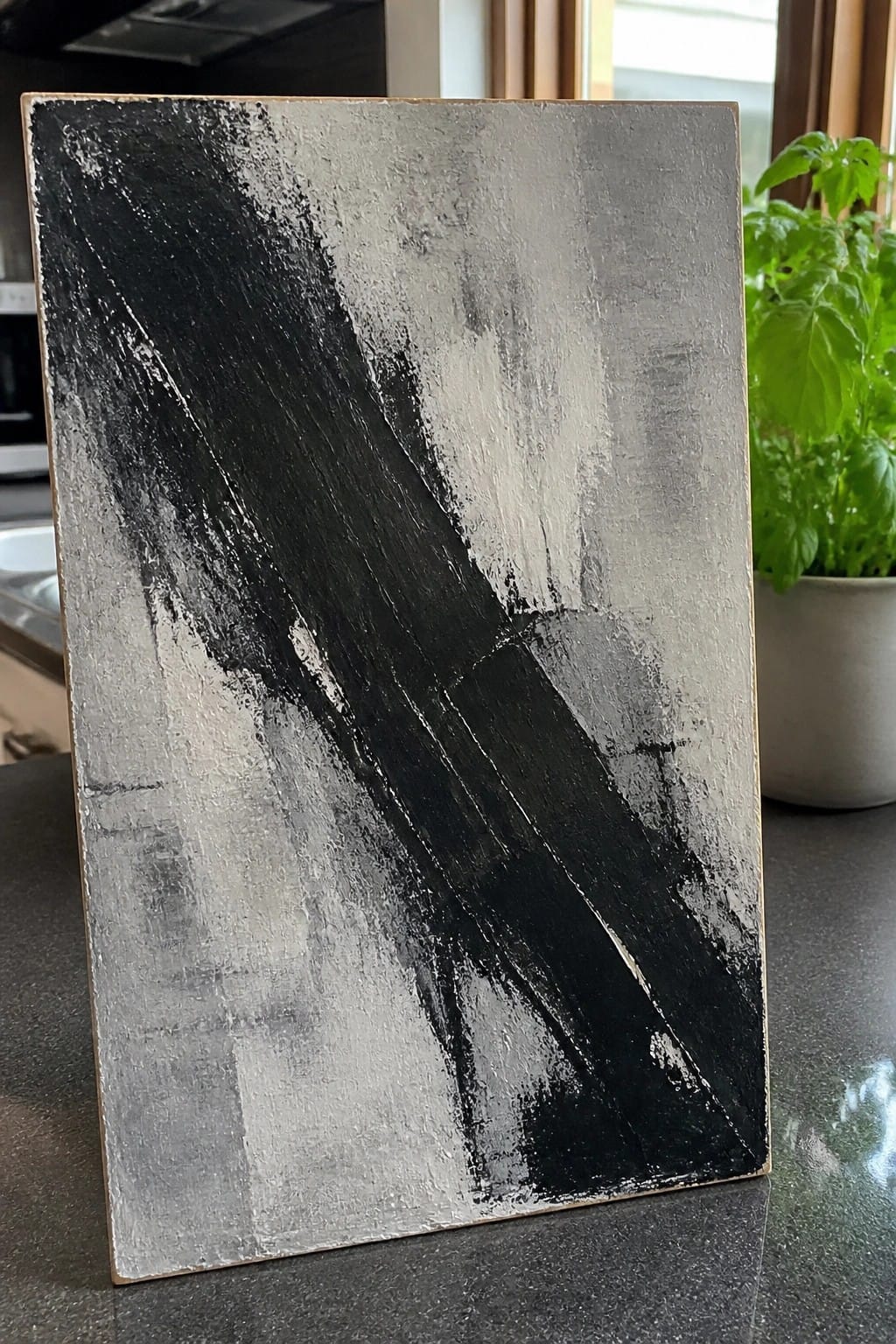 Abstract black and gray painting with bold diagonal strokes on kitchen counter beside potted plant