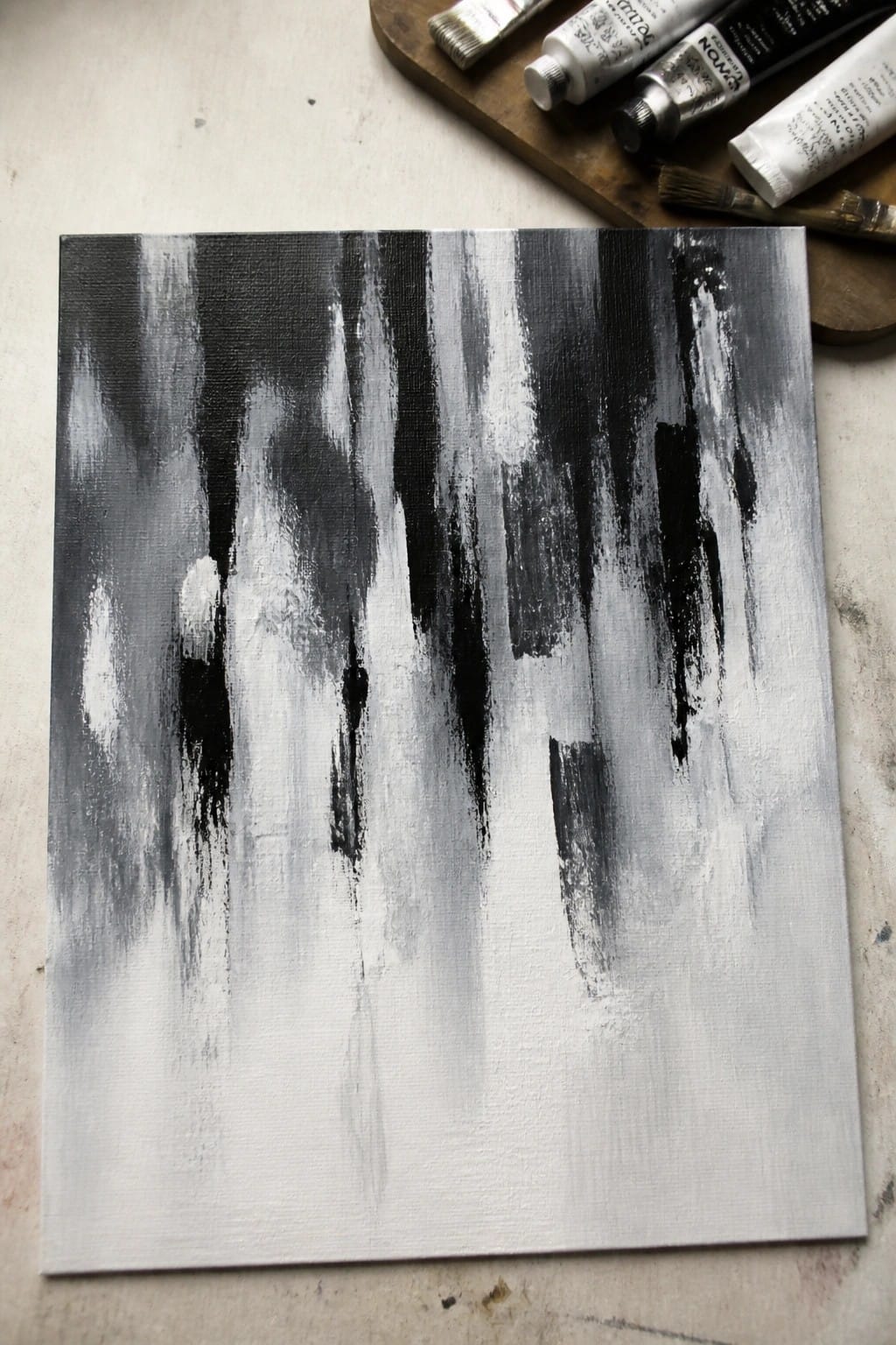 Abstract black and white painting with vertical brushstrokes beside paint tubes and brushes.