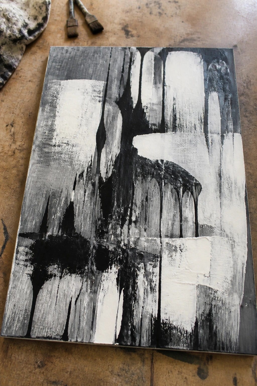 A black and white abstract oil painting on canvas with vertical drips and broad brushstrokes, resting on a wooden surface beside two brushes and a rag.