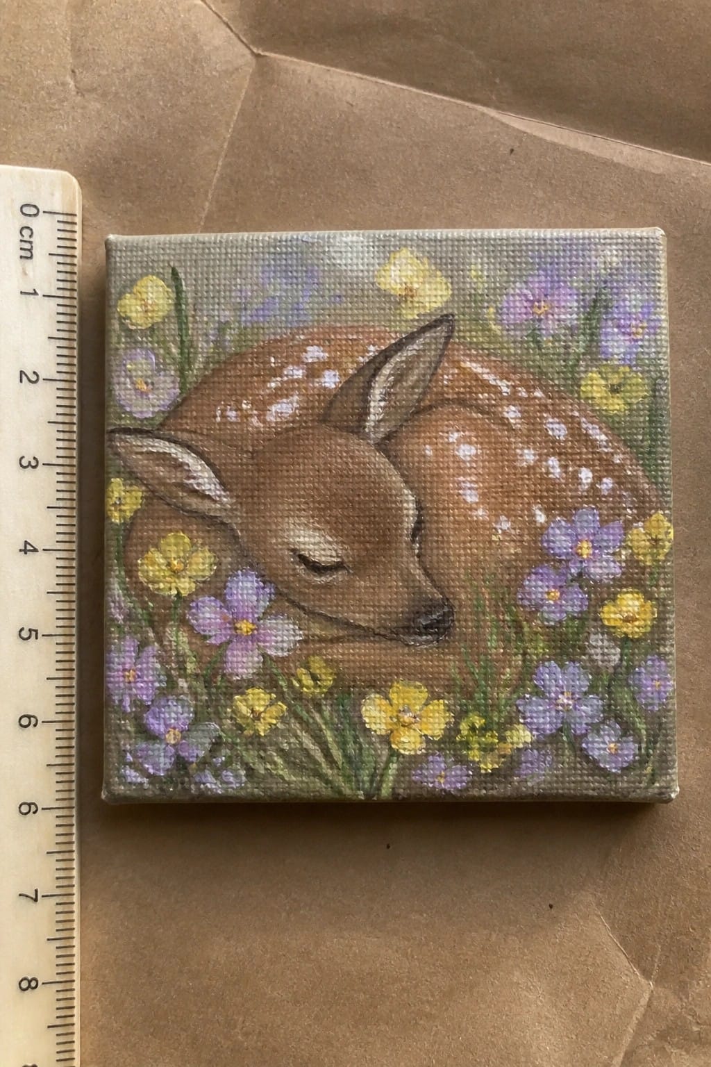 Small square canvas painting of sleeping spotted fawn amid yellow and purple flowers beside ruler.