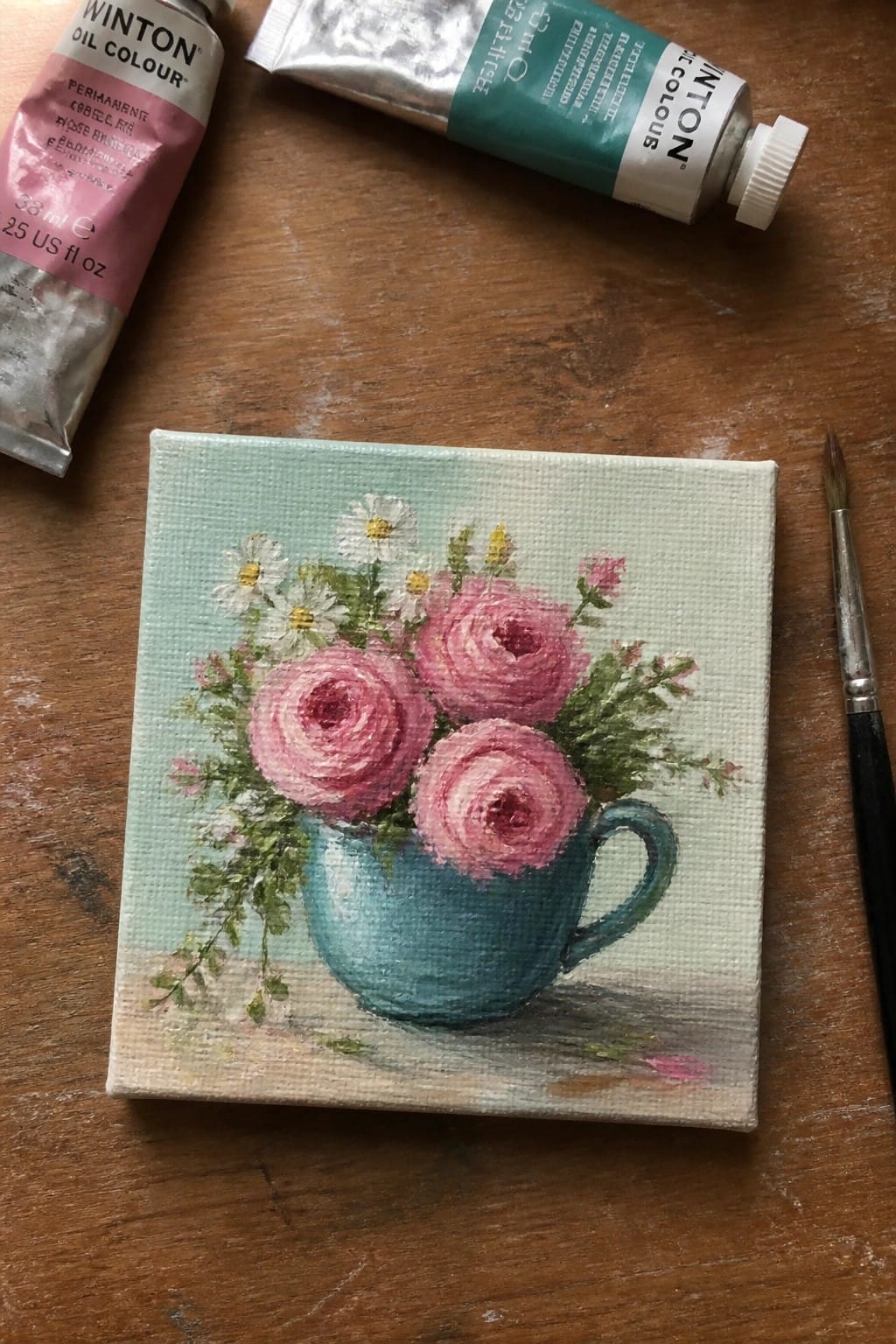 Small floral oil painting of pink roses in blue cup with paint tubes and brush on wood