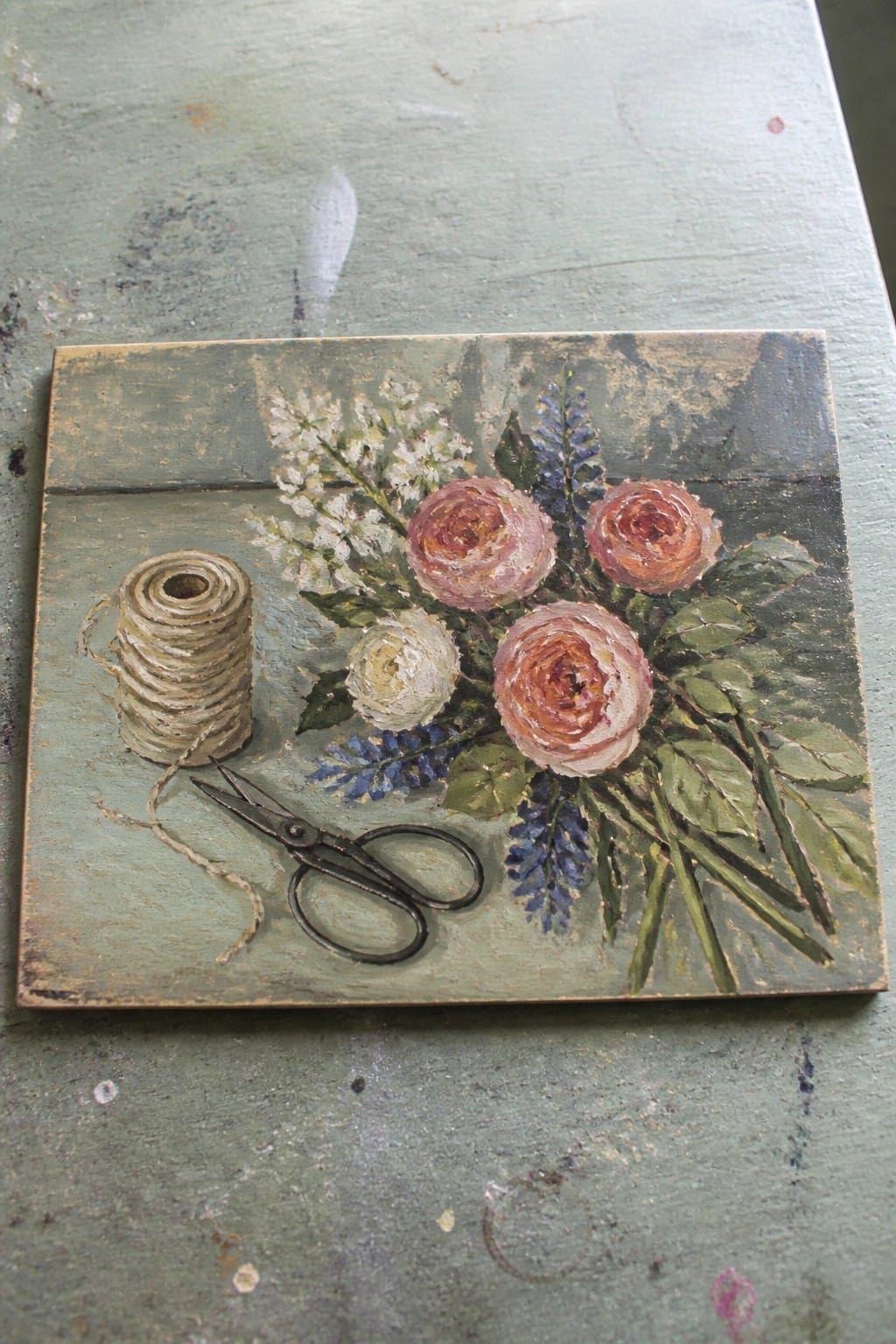 An oil painting of pink roses and wildflowers beside a spool of twine and scissors on a green wooden surface.