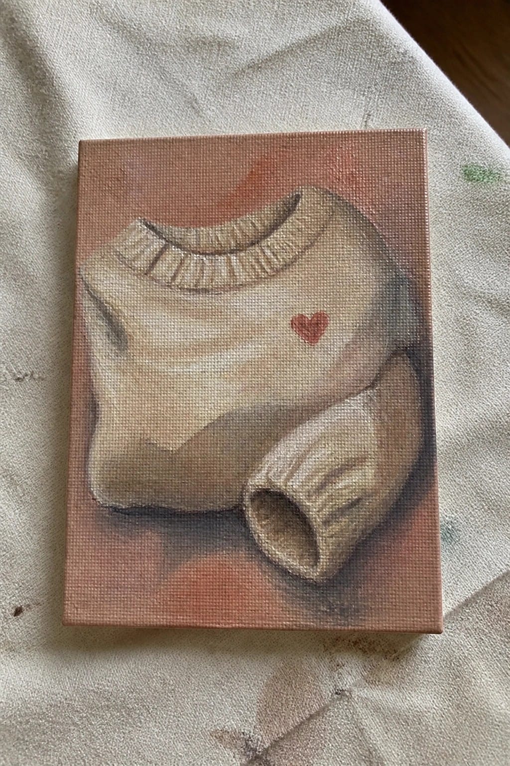Beige turtleneck sweater with red heart painted on small canvas over white cloth.