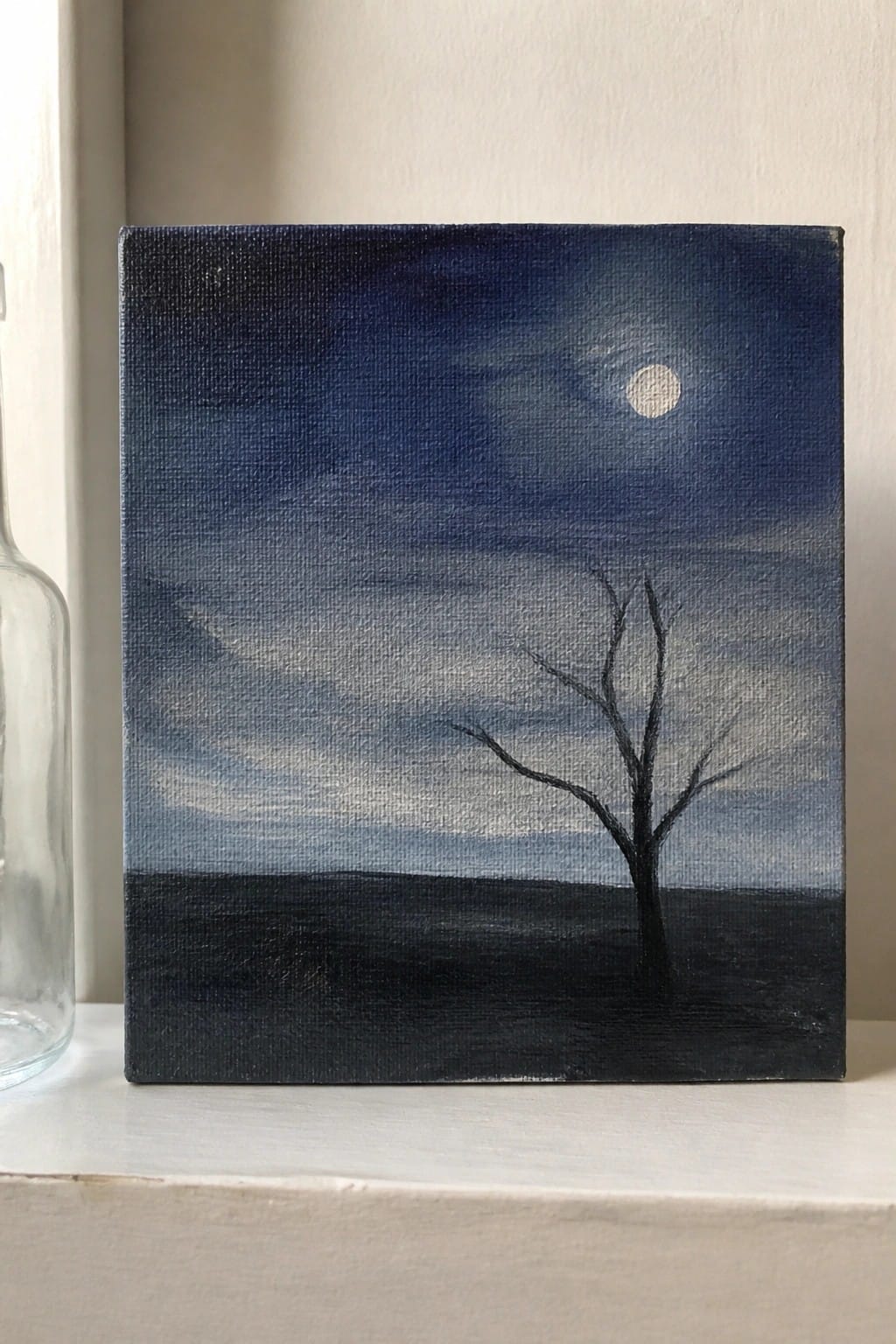 Dark blue night sky painting with moon and bare tree on canvas next to bottle