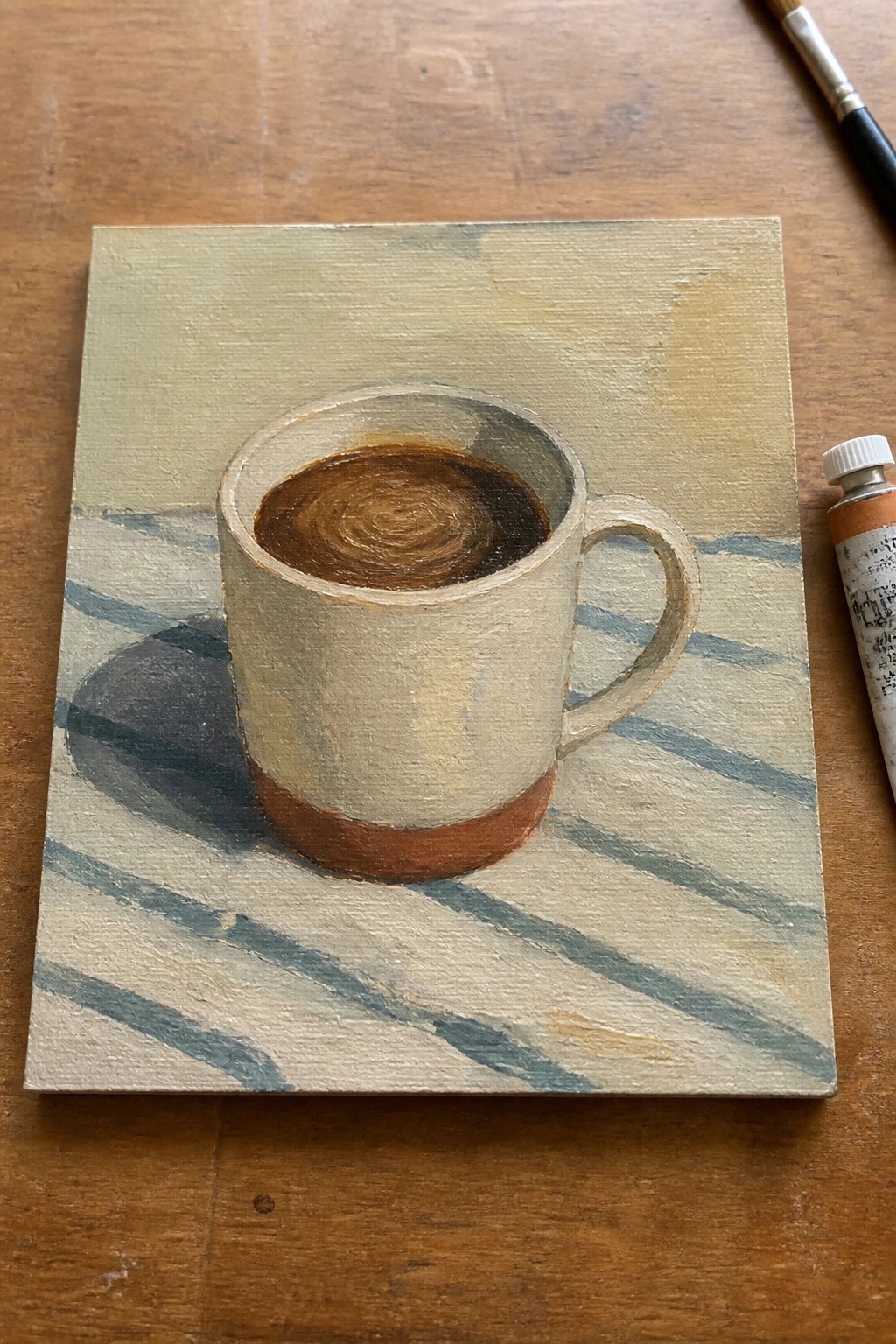 Oil painting of coffee mug on blue-striped cloth beside paint tube and brush