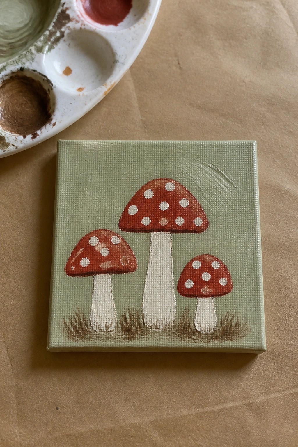 Three red spotted mushrooms with white stems on small green canvas beside palette