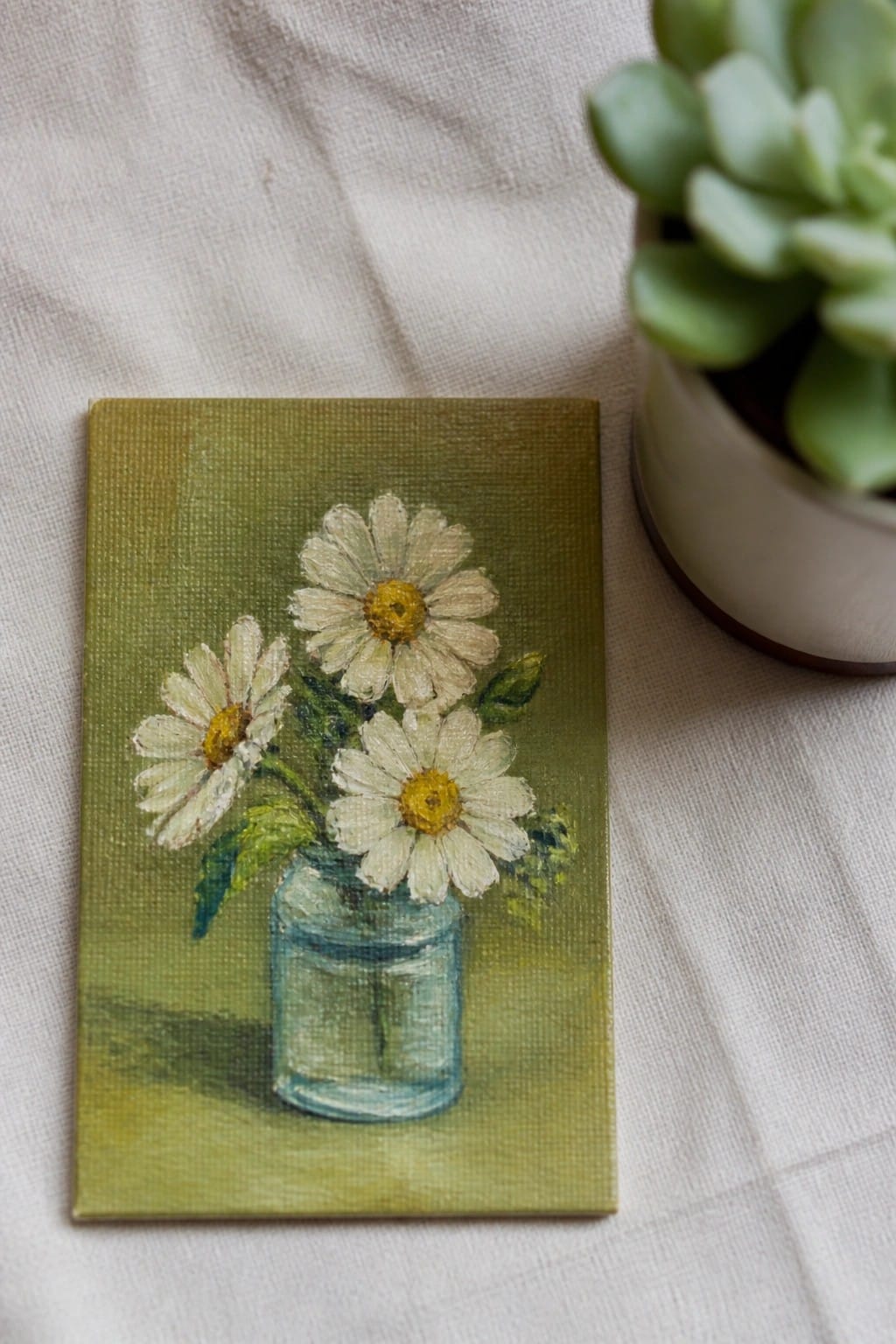 Small canvas painting of three white daisies in blue glass jar beside succulent