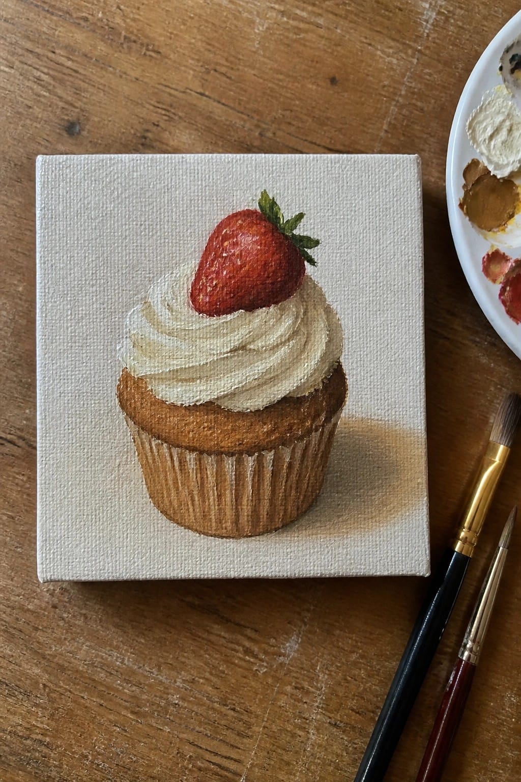 Realistic strawberry cupcake painting on white canvas with palette and brushes on wood.