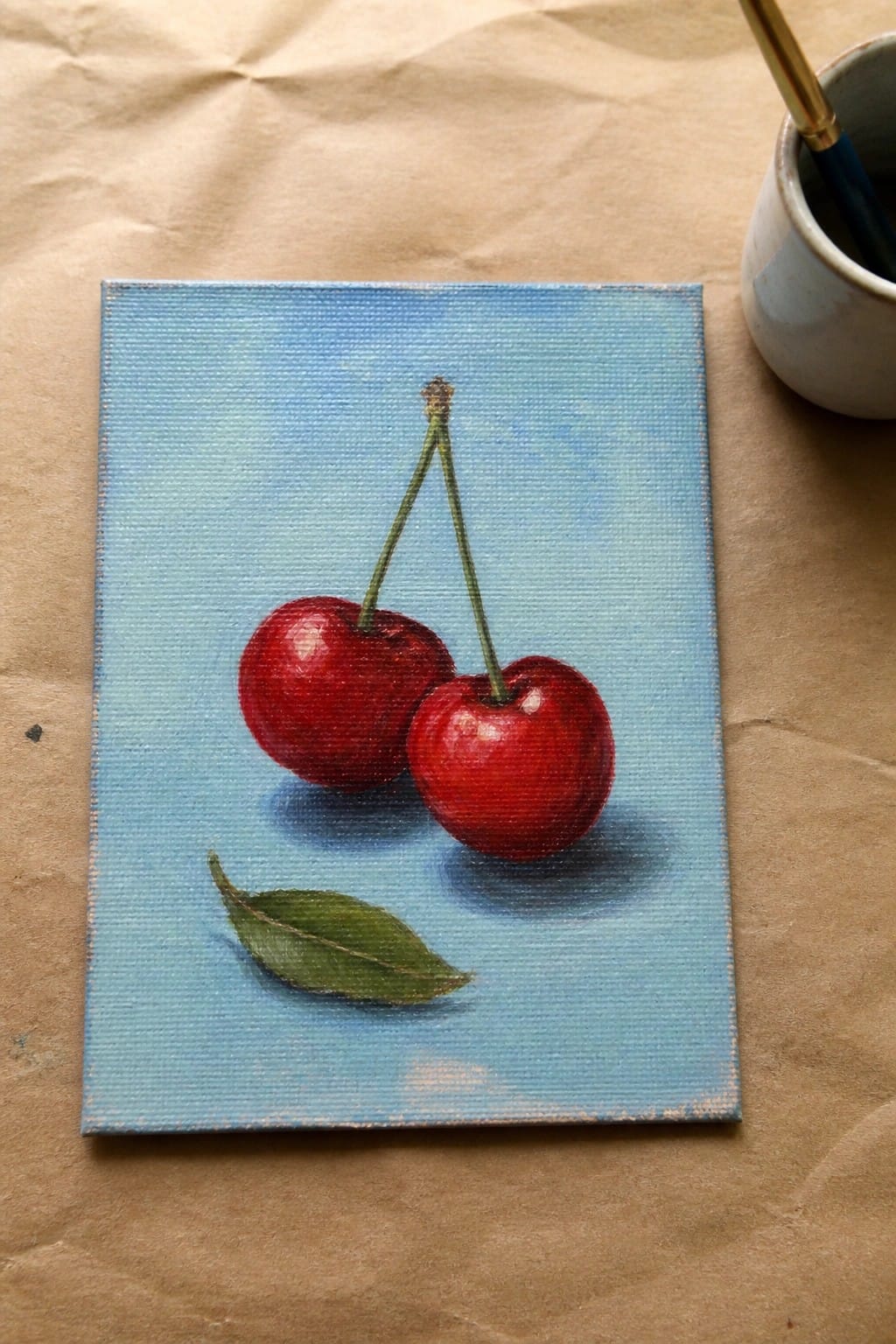 Realistic painting of two red cherries with green stems and leaf on light blue canvas beside paintbrush.