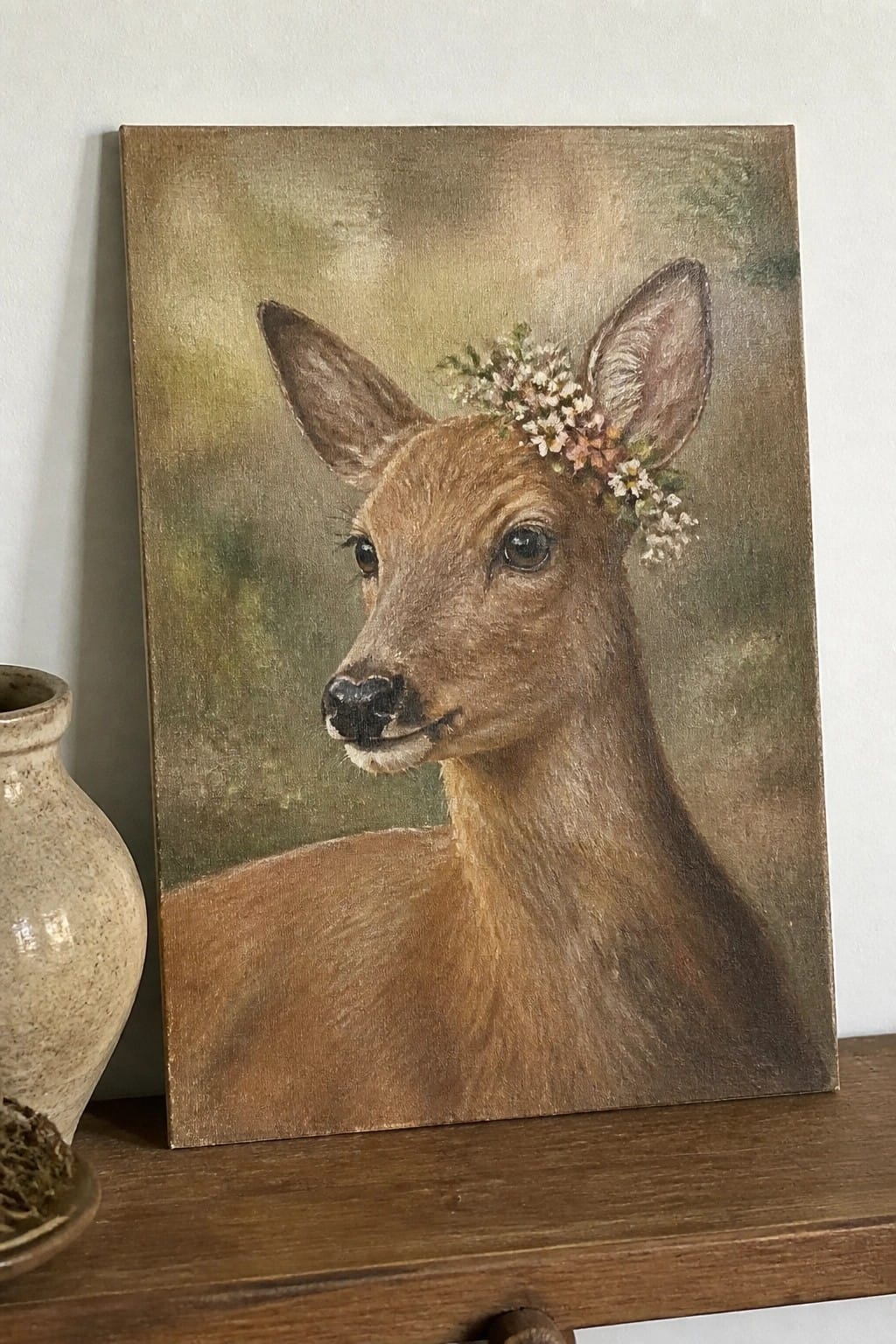 A realistic oil painting of a brown deer with a flower crown of white and pink blossoms on its head, shown in profile view against a soft green-brown background.