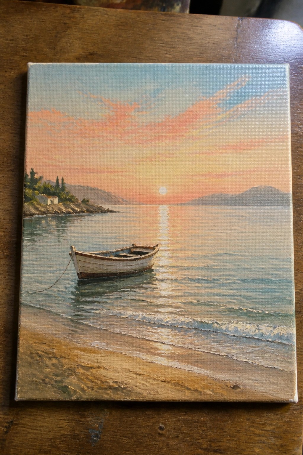 Small boat on sandy beach at sunset with sun reflecting on calm sea and hills.