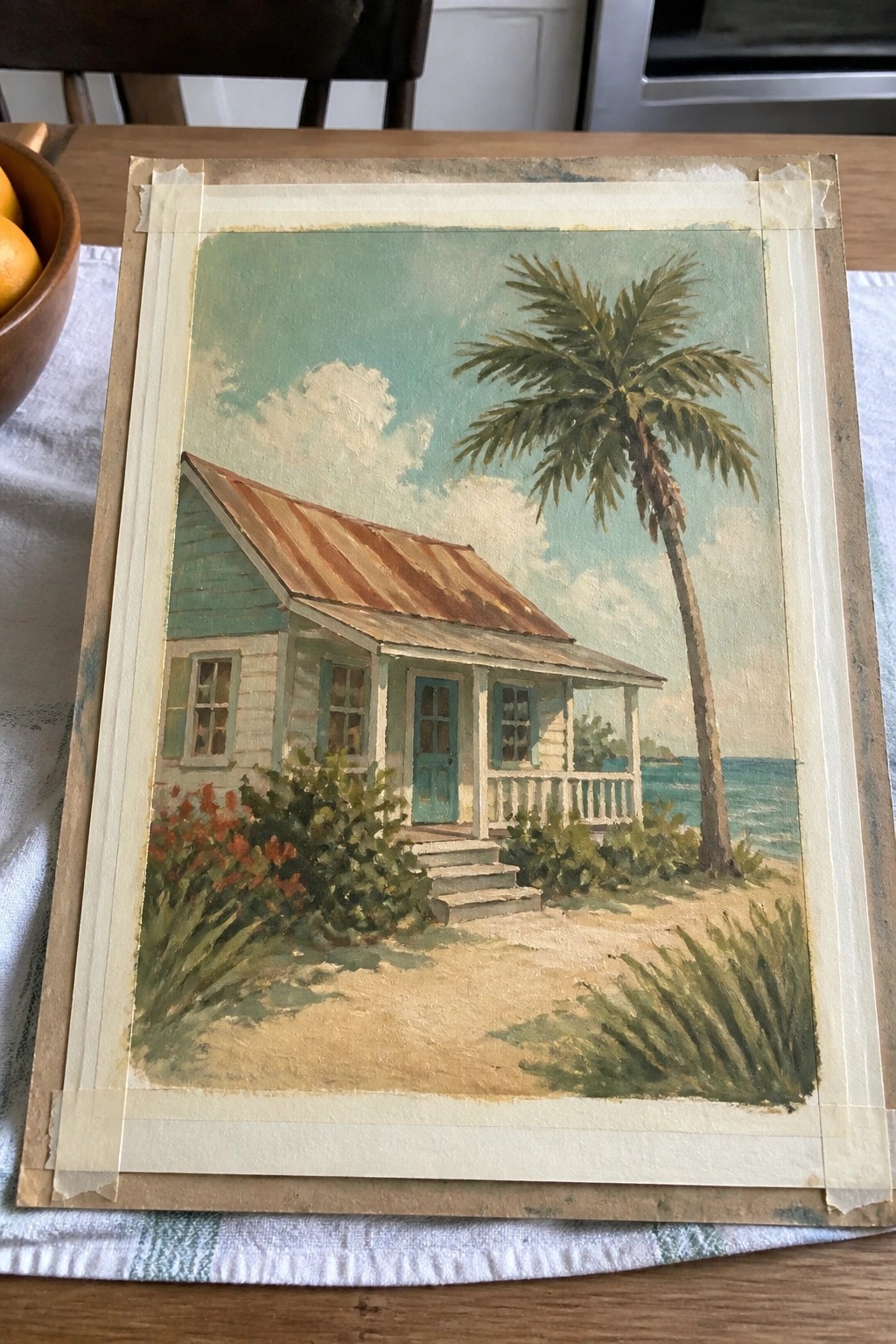 Painting of turquoise beach cottage with palm tree and ocean, taped to board on table