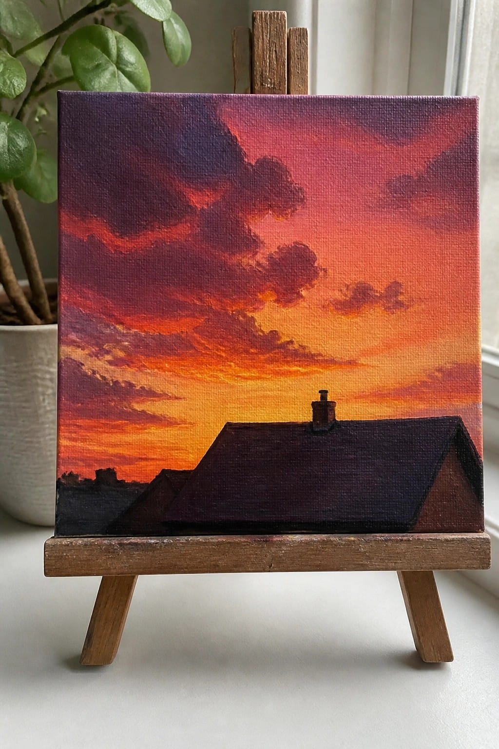 Canvas on wooden easel shows vibrant red-orange sunset over dark house silhouette.
