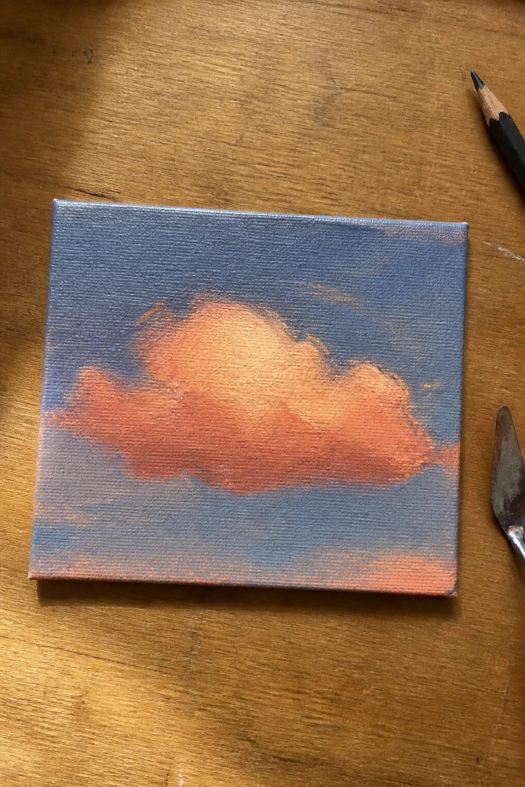 Small canvas with orange clouds on blue sky beside pencil and palette knife on wood