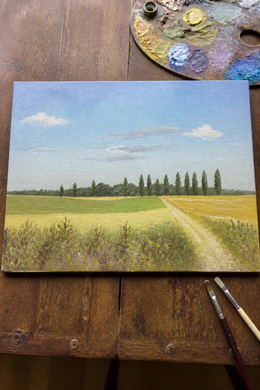 Canvas landscape of fields and trees with palette and brushes on wooden table