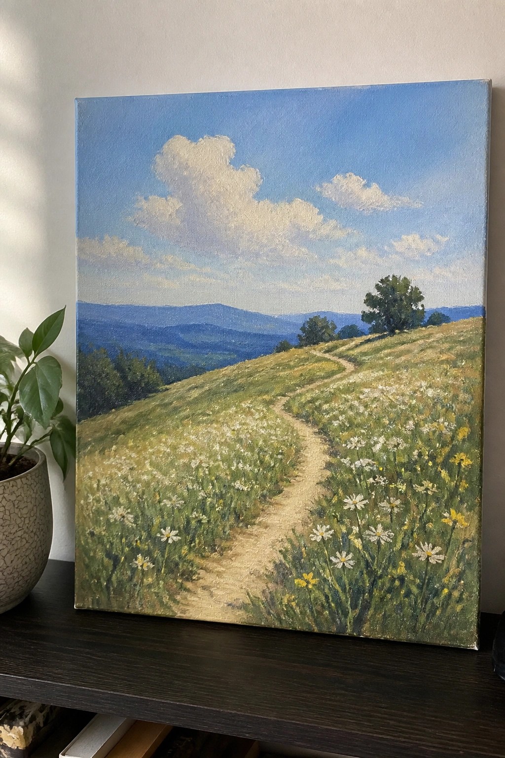 Landscape painting of flowery hillside path with mountains beside potted plant on shelf.