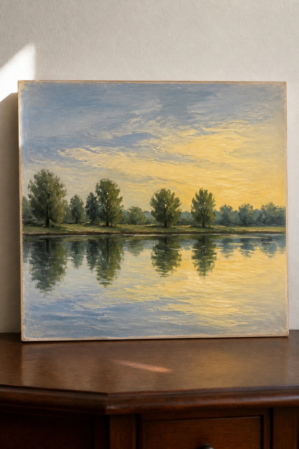 An oil painting of a calm body of water with trees along the far shore and their reflections under a blue and yellow sky, resting on a wooden surface.