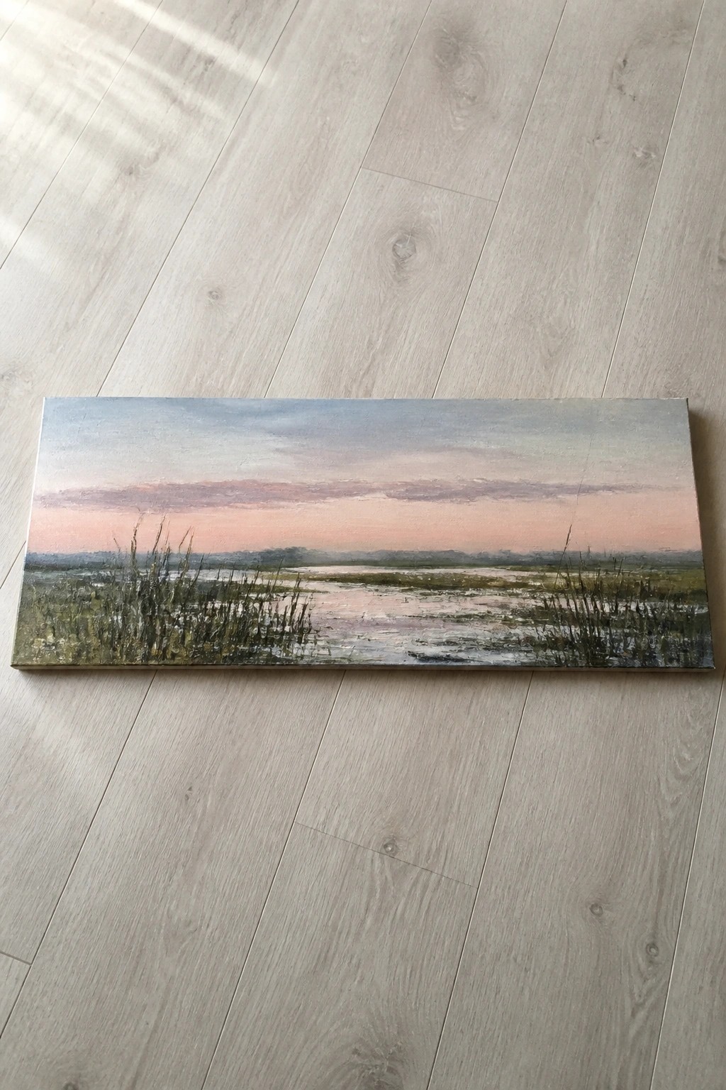 Wide landscape painting of pink sunset over marsh with reeds on wooden floor