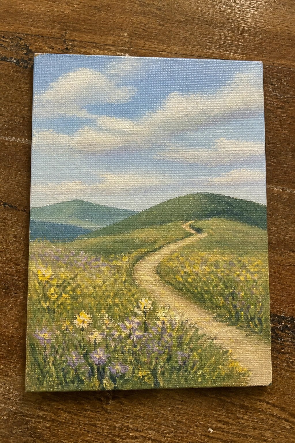 Canvas painting of winding path through yellow-purple wildflowers with green hills and blue sky