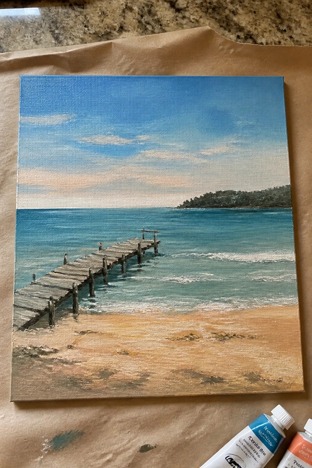 Painting of wooden pier on sandy beach with turquoise ocean, blue sky, paint tubes nearby.