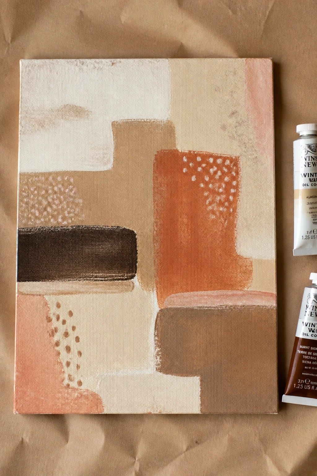Abstract canvas with geometric shapes in earth tones beside two paint tubes on brown paper
