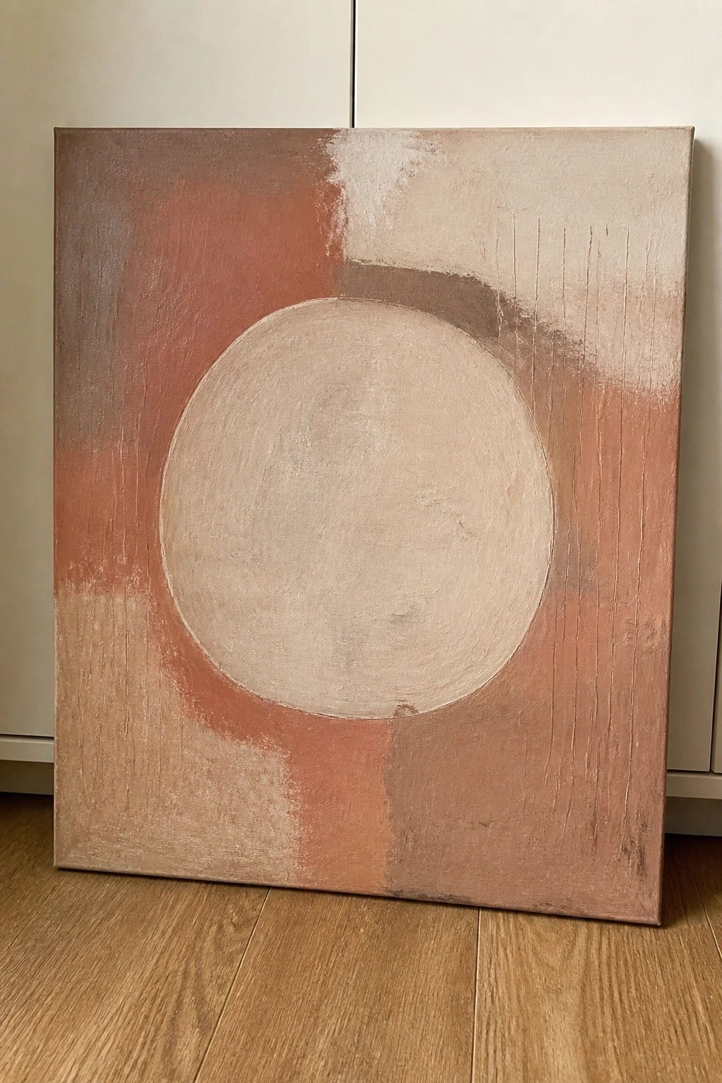 Abstract painting with large beige circle on textured terracotta background, leaning on wood floor