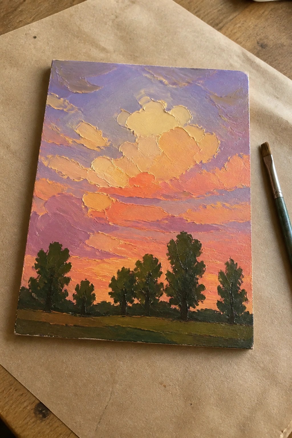 Vibrant impasto sunset painting with purple-orange clouds above green trees and a paintbrush