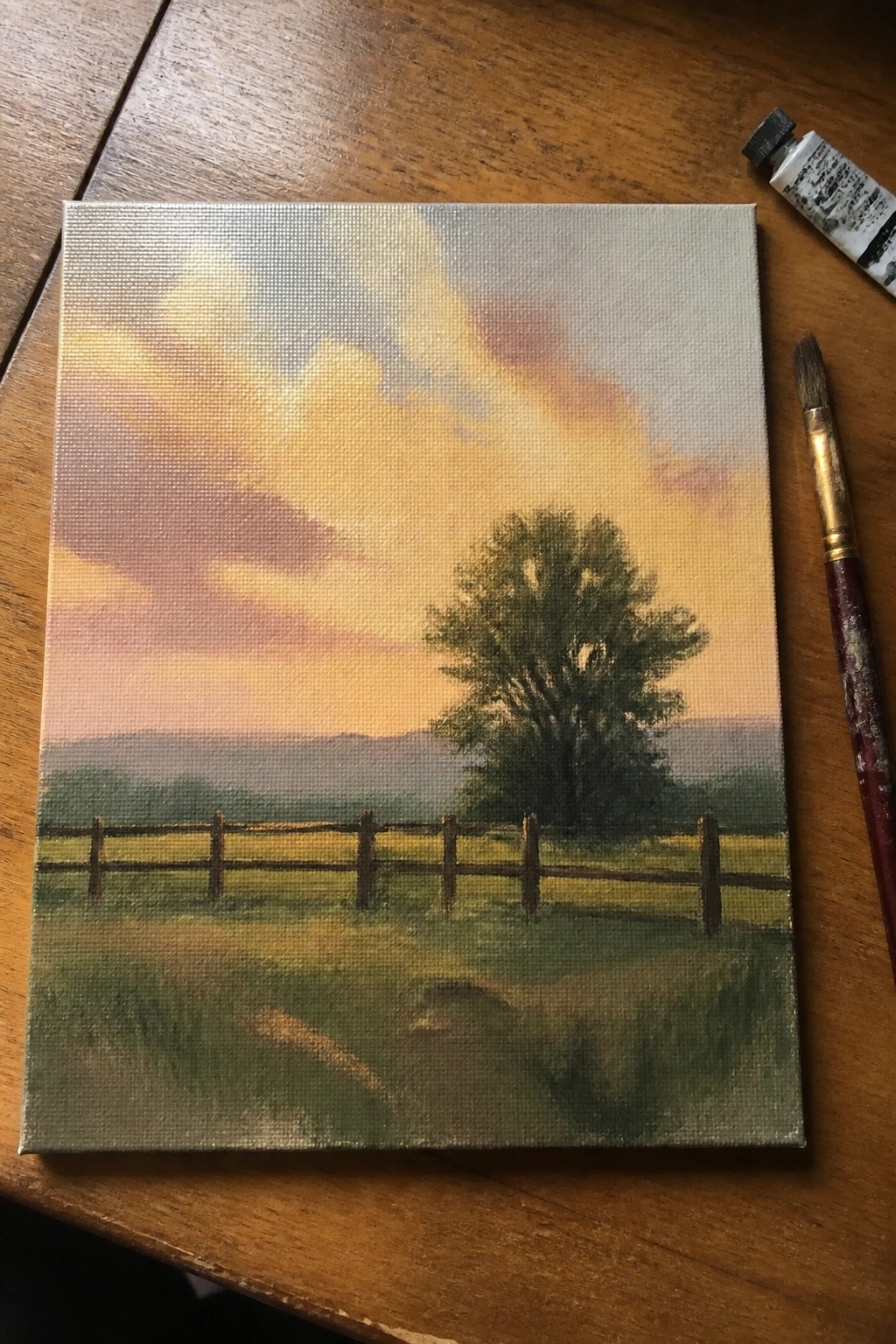 Sunset landscape painting on canvas with fence, tree, beside paintbrush and tube on wood.
