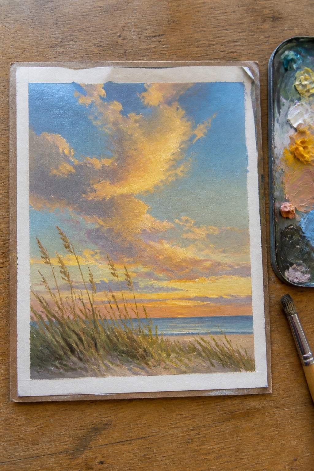 Sunset seascape painting with golden clouds, ocean, grasses, palette and brush on wood.