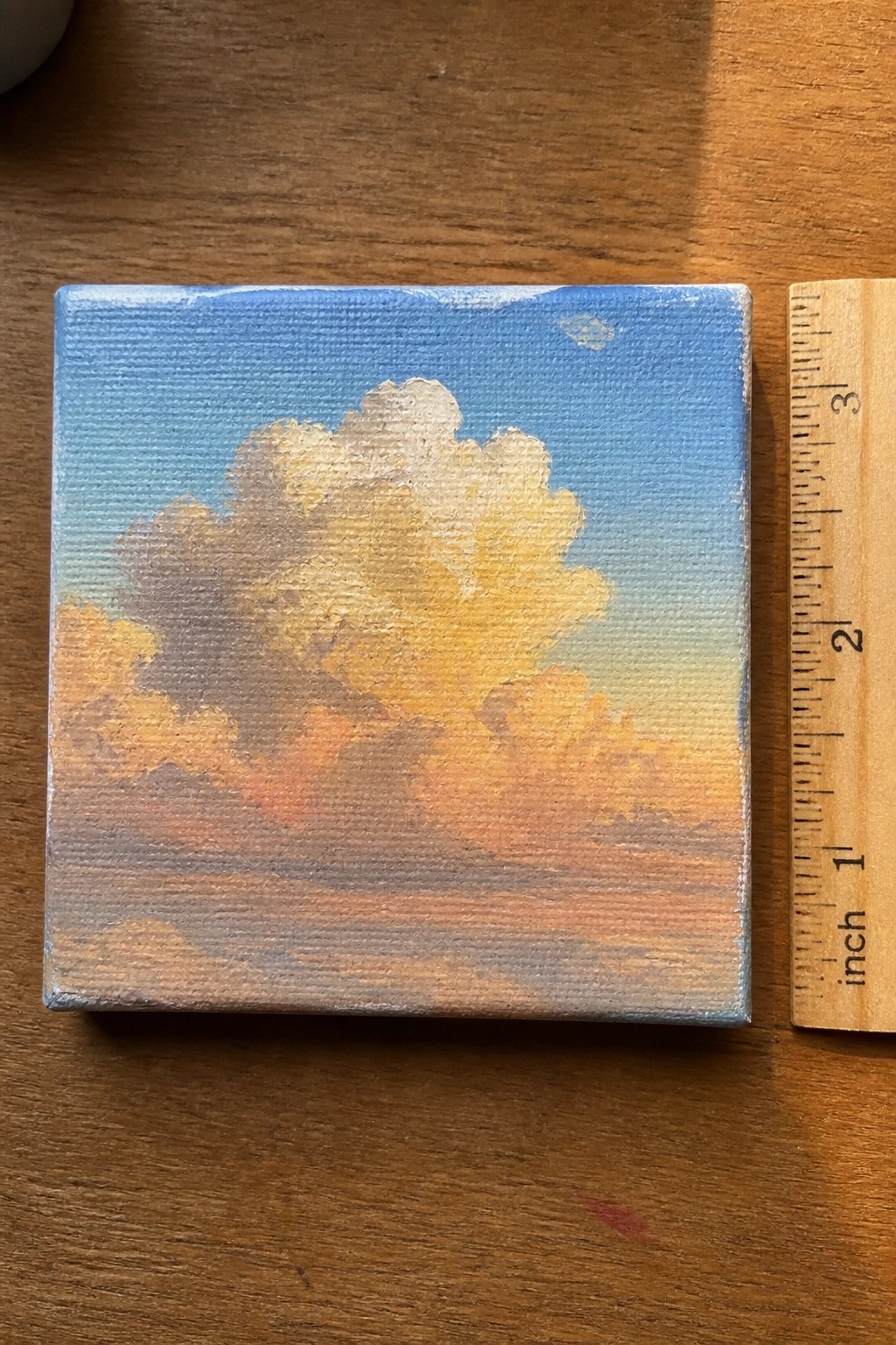 Small square oil painting of a golden hour cloud in yellow and orange tones against a blue sky next to a wooden ruler.