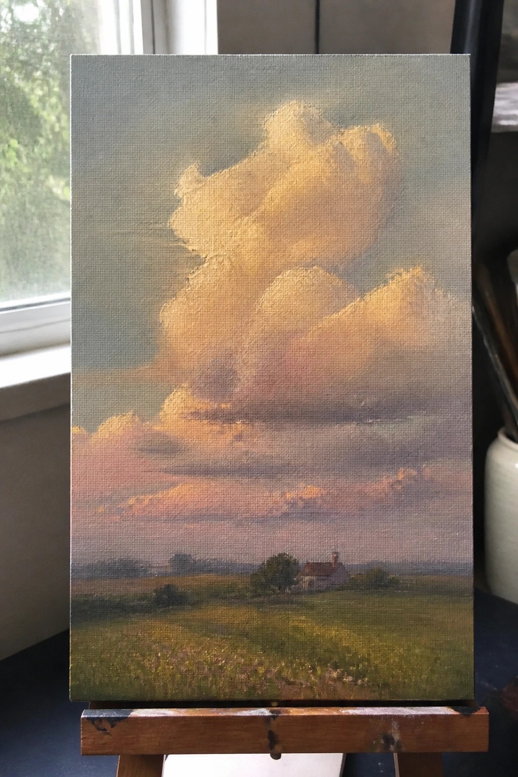 Realistic oil painting on easel of golden-pink clouds over green field with distant house.
