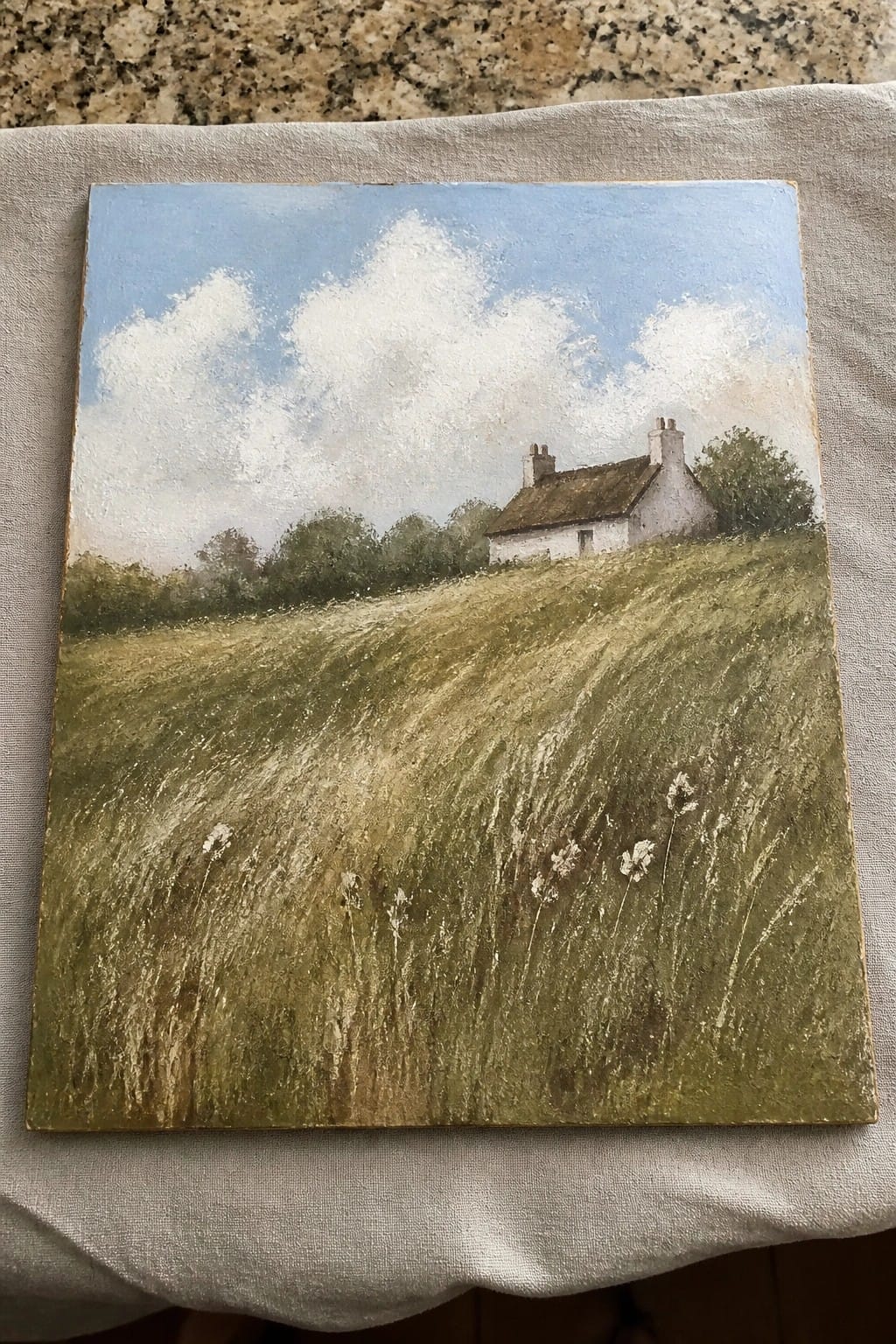 Landscape painting of white cottage on grassy hill under cloudy blue sky, resting on cloth