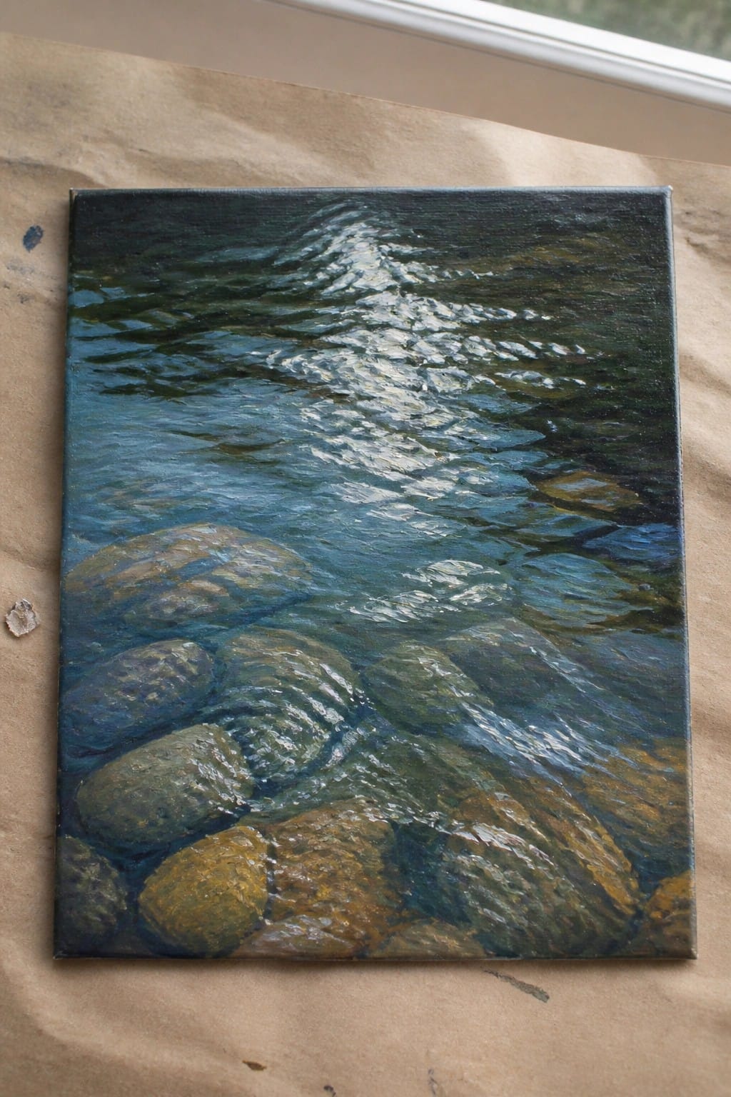 Rectangular canvas painting of rippling blue water over multicolored rocks with sunlight reflections