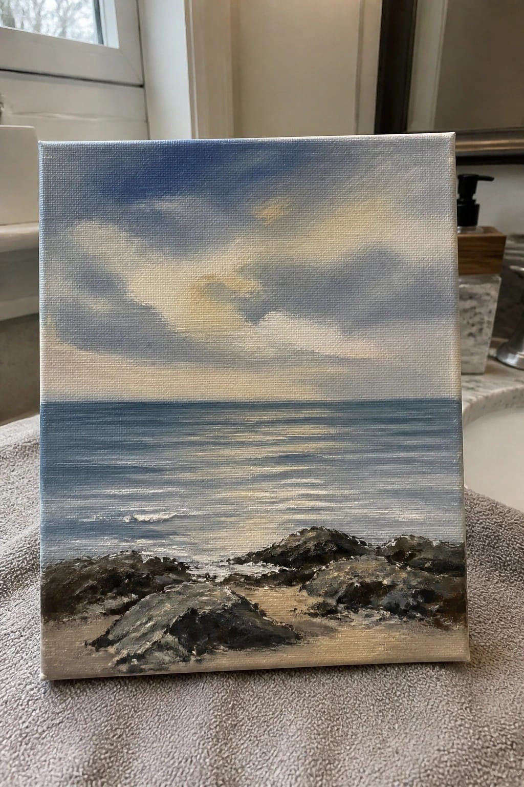 Seascape painting on canvas with blue sky, ocean, and dark rocks resting on gray towel.