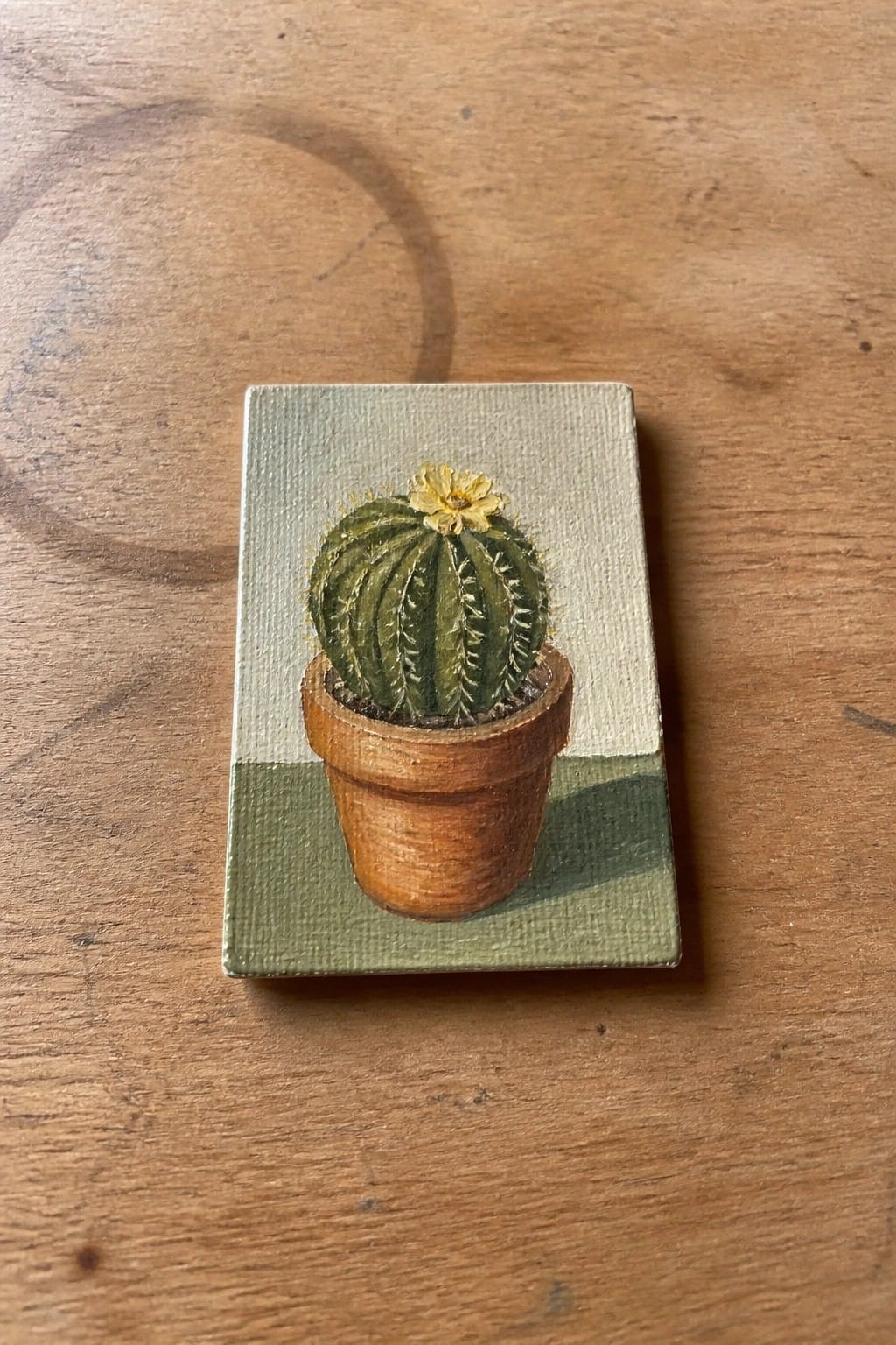 Oil painting of a green cactus with a yellow flower in a brown pot on a light green and dark green background on a small canvas.