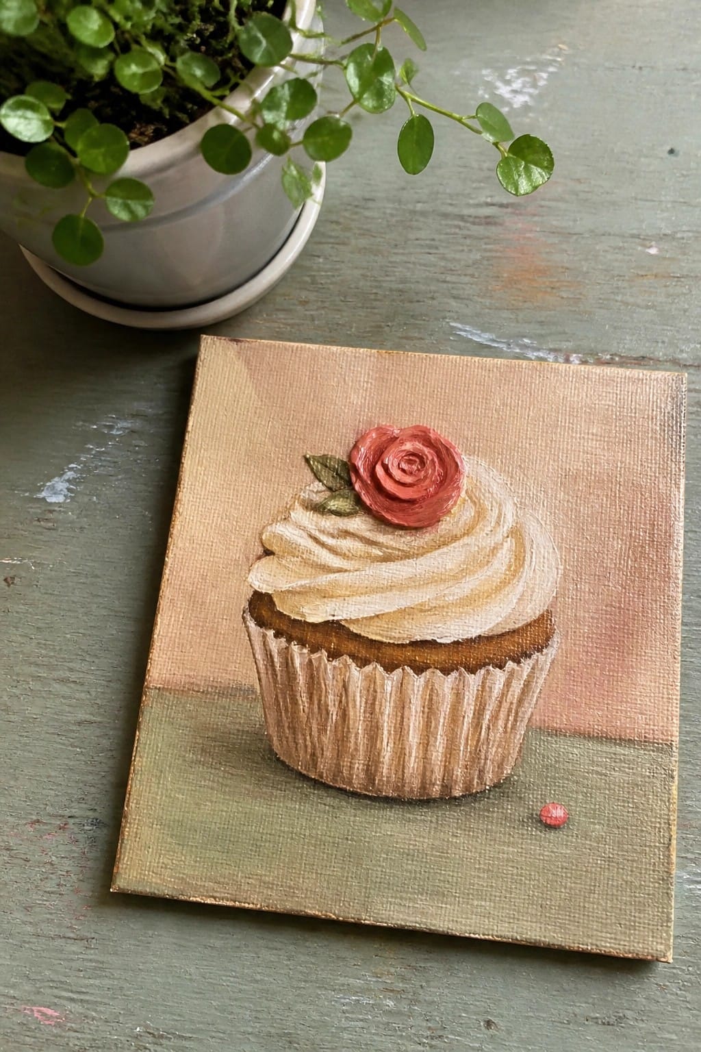 Square canvas painting of cupcake with white frosting and red rose on wooden table
