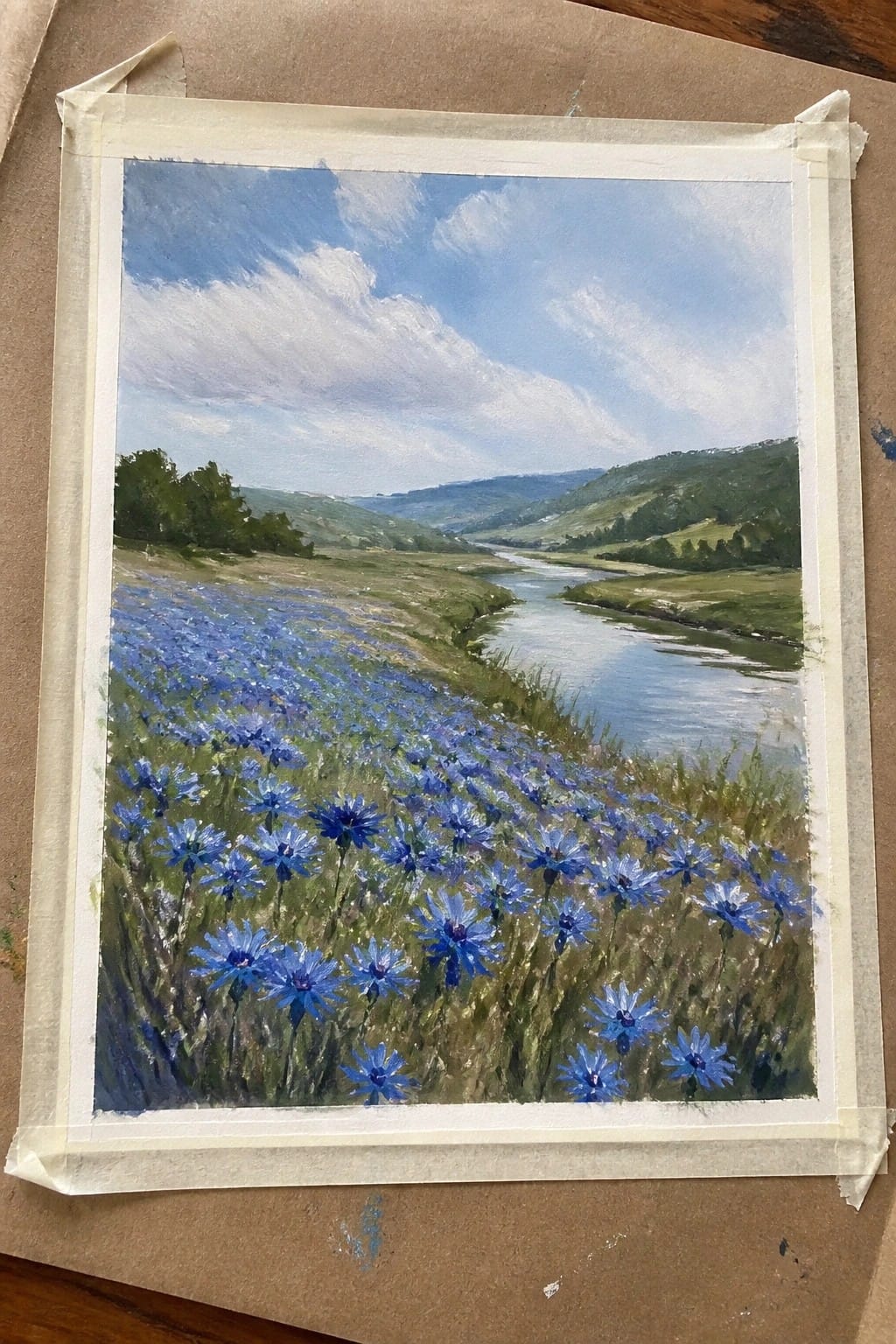 Painted landscape of blue wildflowers along a river winding through green hills under clouds