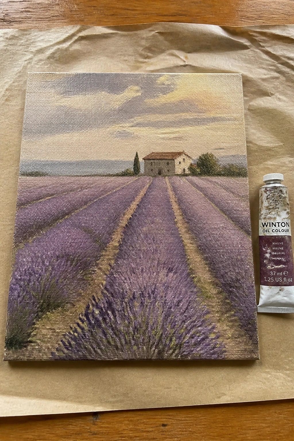 An oil painting of lavender rows leading to a house sits on brown paper beside a tube of mauve oil paint.