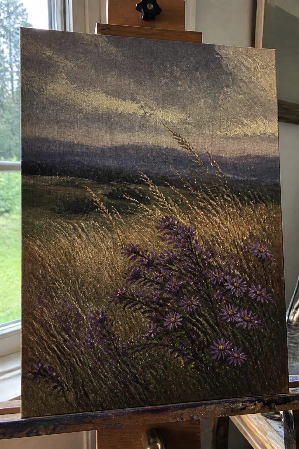 Oil painting on easel depicts purple wildflowers amid golden grasses under stormy skies.