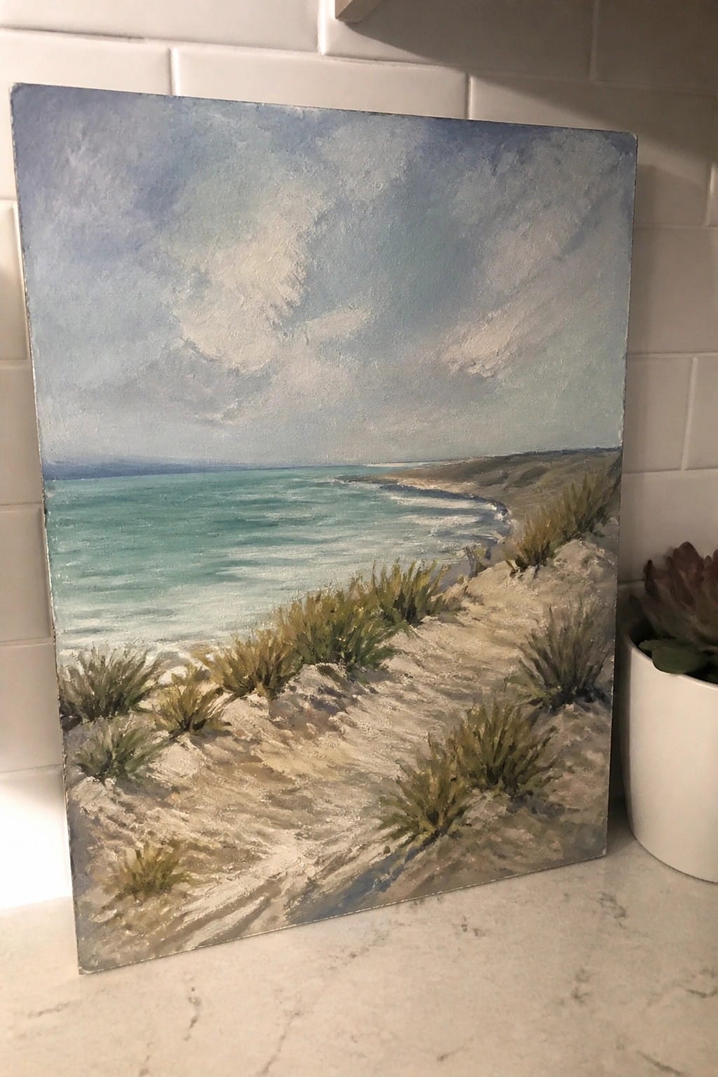 Beach painting with grassy dunes and turquoise sea beside potted succulent on tiled counter.