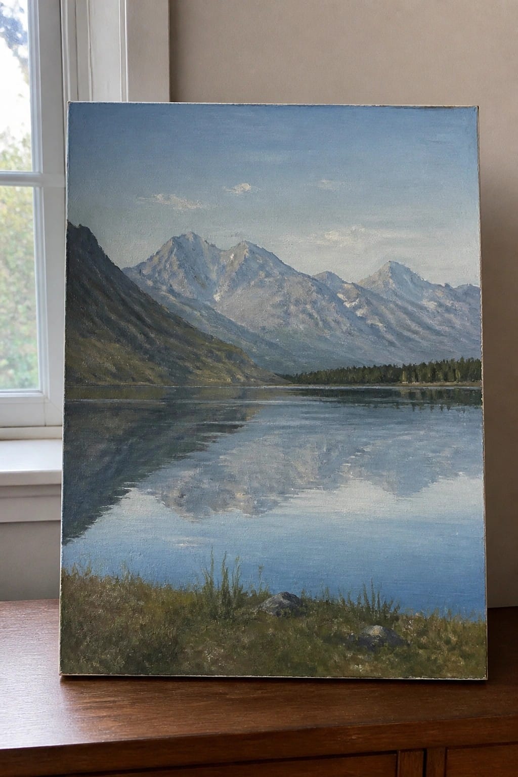 A realistic oil painting of a calm blue lake reflecting a range of mountains under a pale sky, with grass and rocks in the foreground.