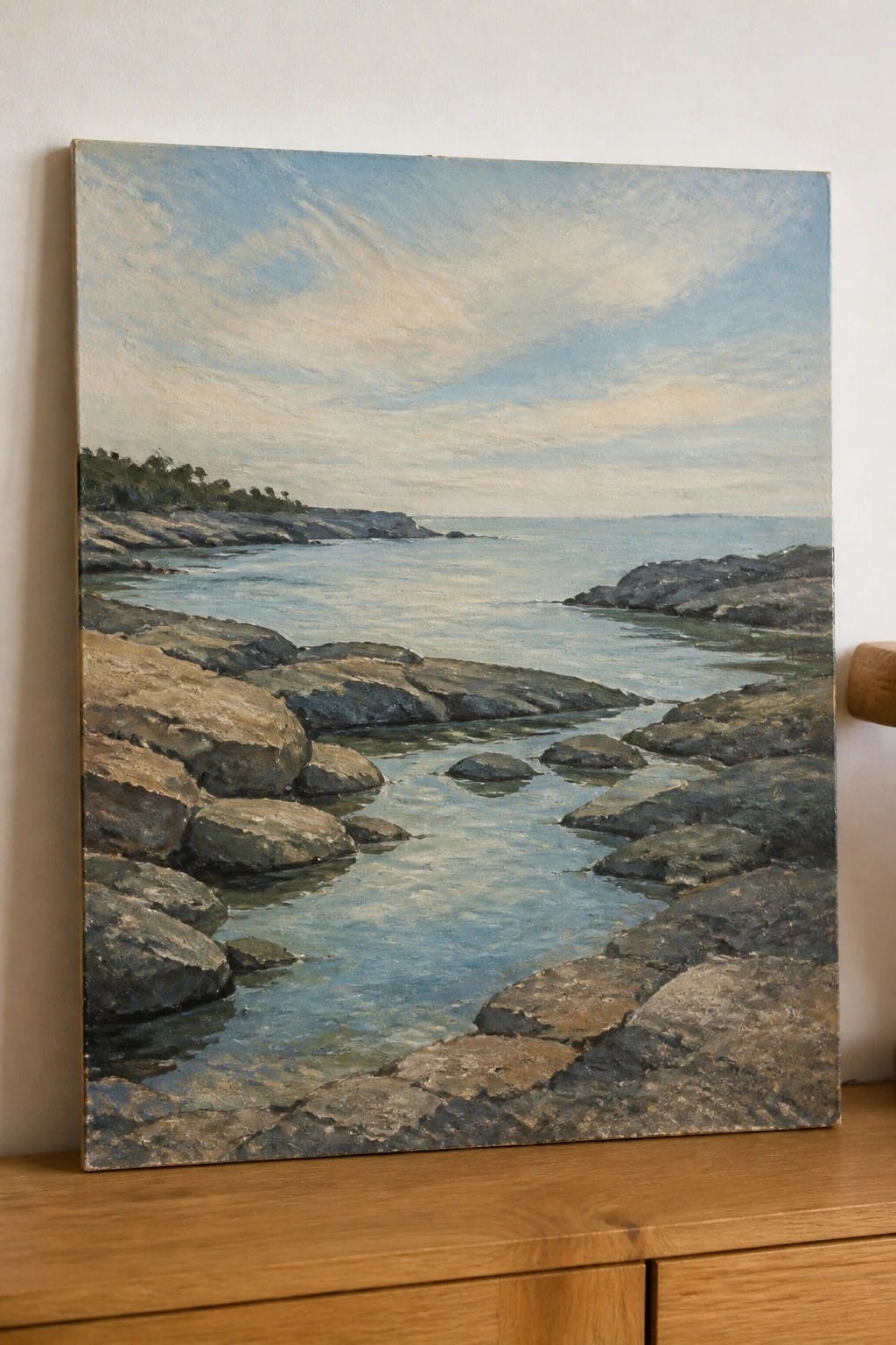 Framed oil painting of rocky coastline with calm blue water under cloudy sky on wood surface