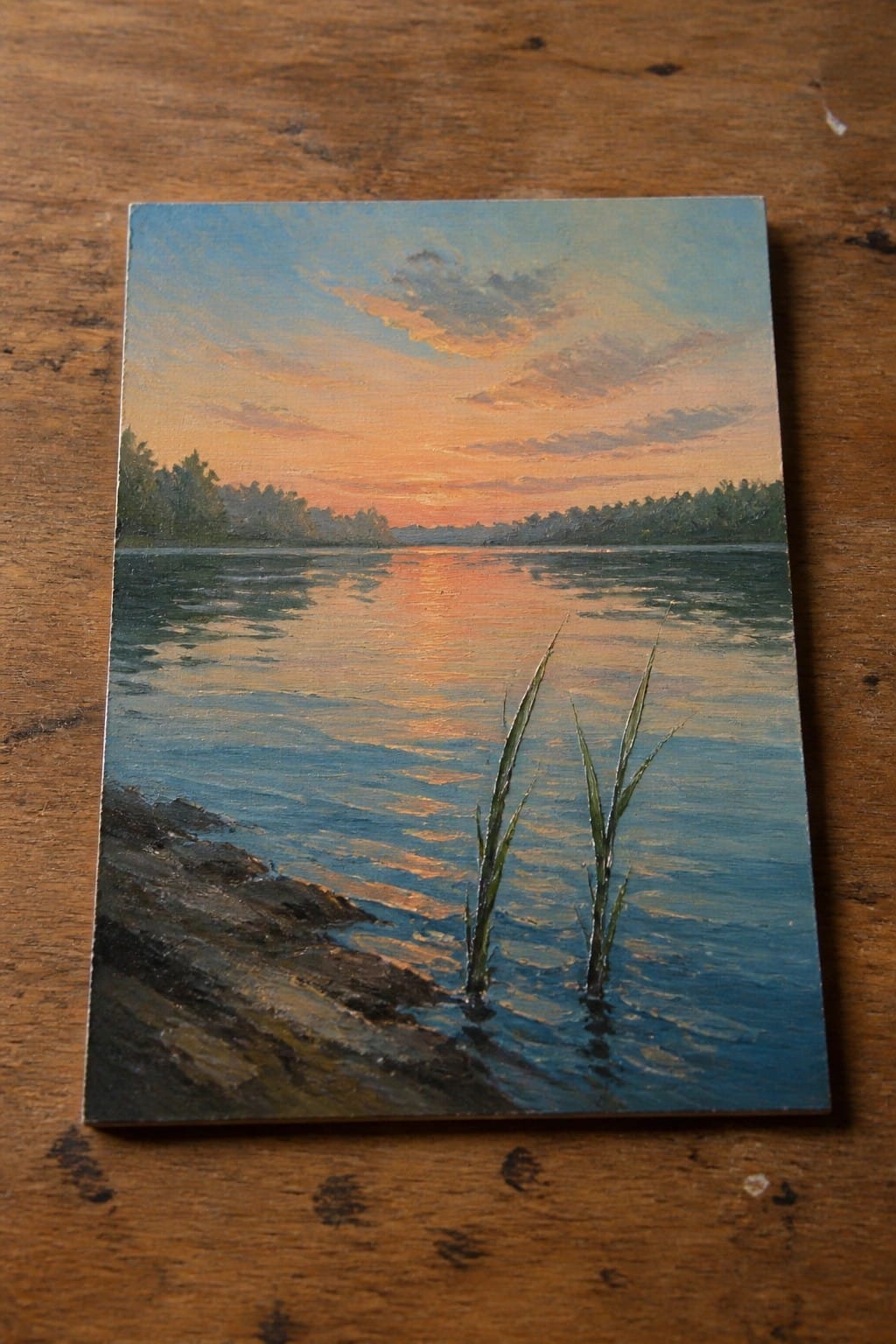 An oil painting of a sunset over a lake with reeds growing from the water near a rocky shore and trees on the far bank.