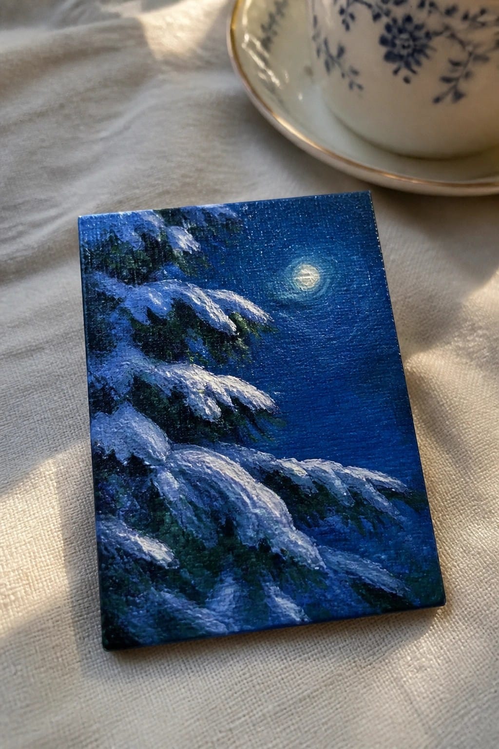 Small rectangular painting of snow-covered pine trees under a glowing moon beside floral teacup