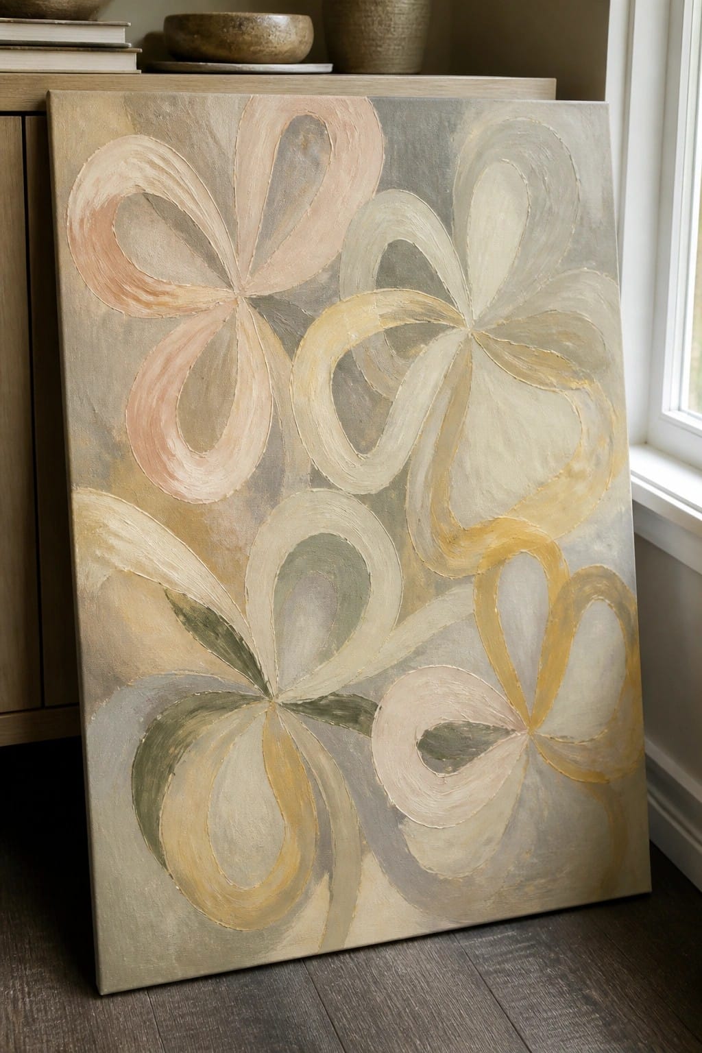 An abstract oil painting of large overlapping looped petal shapes in soft pink, beige, yellow, and green on a muted gray background, leaning against a wooden cabinet.
