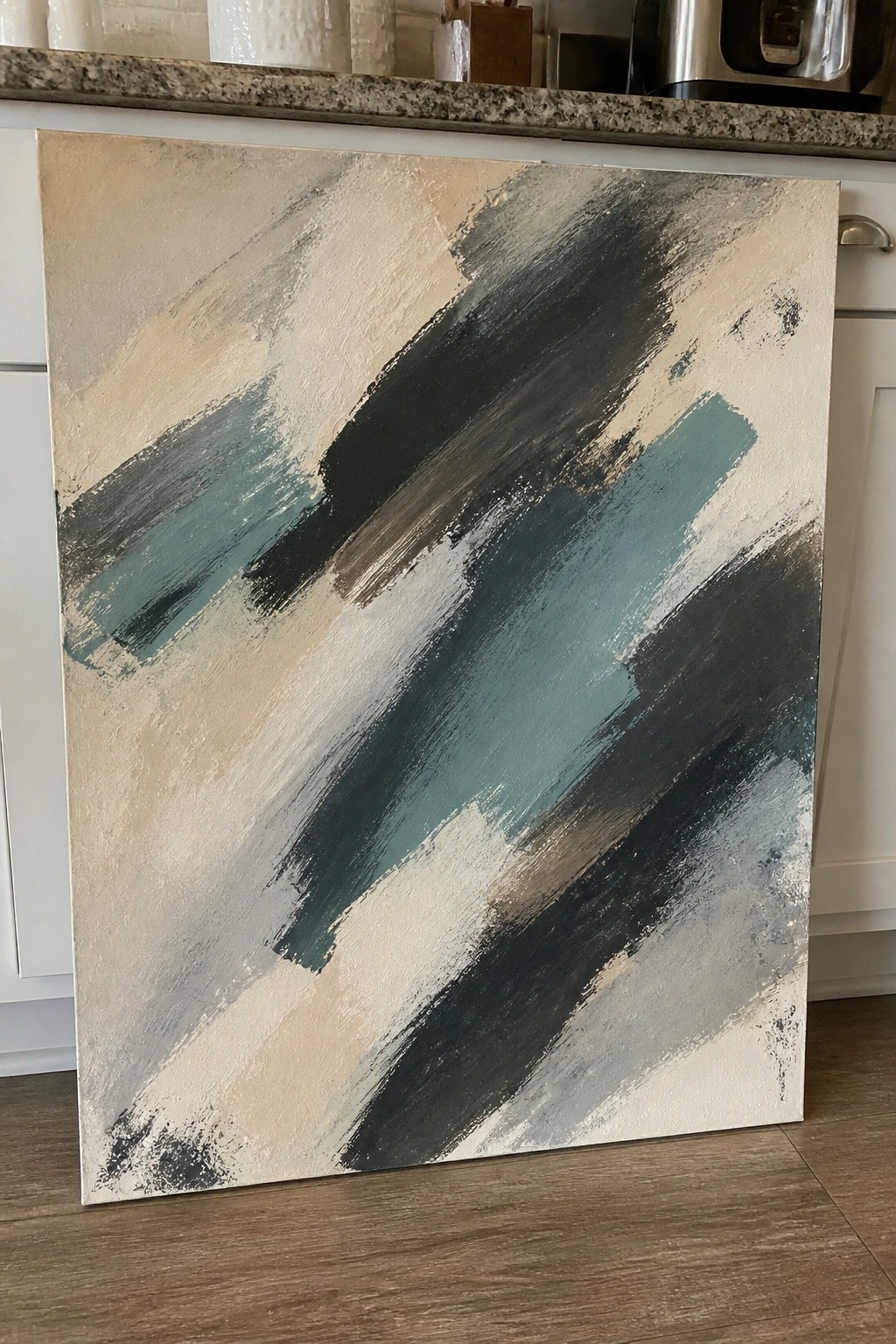 Abstract painting with diagonal teal, black, and beige brushstrokes leaning against kitchen cabinets