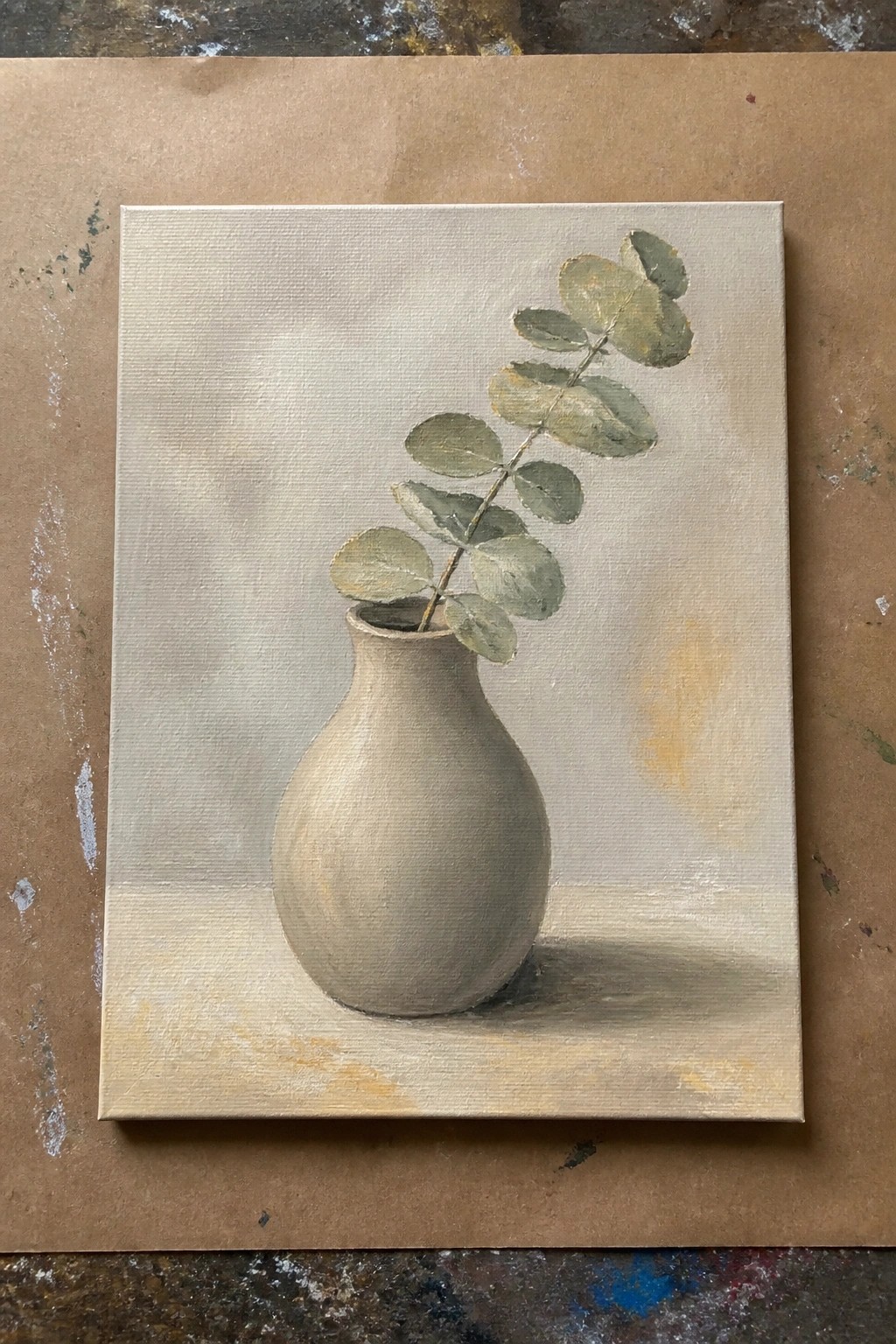 A realistic oil painting of a beige vase holding a single eucalyptus branch against a light neutral background, with soft shadows on the surface.
