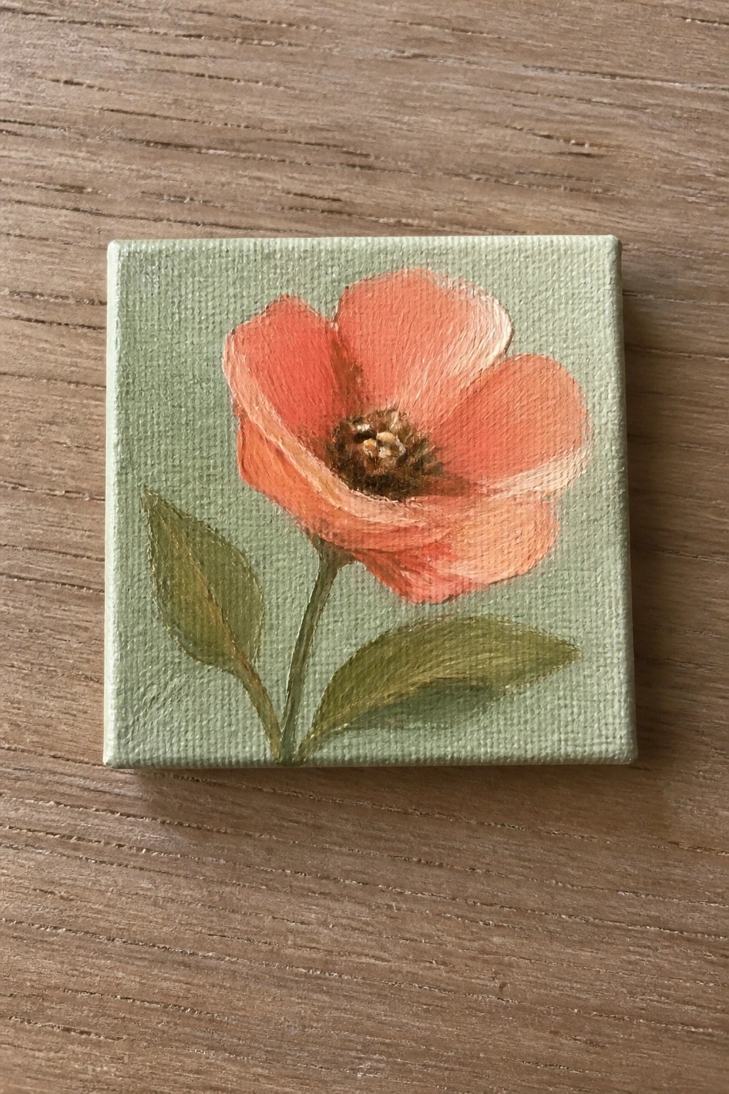 Small square canvas with coral flower, green stem and leaves on wood table