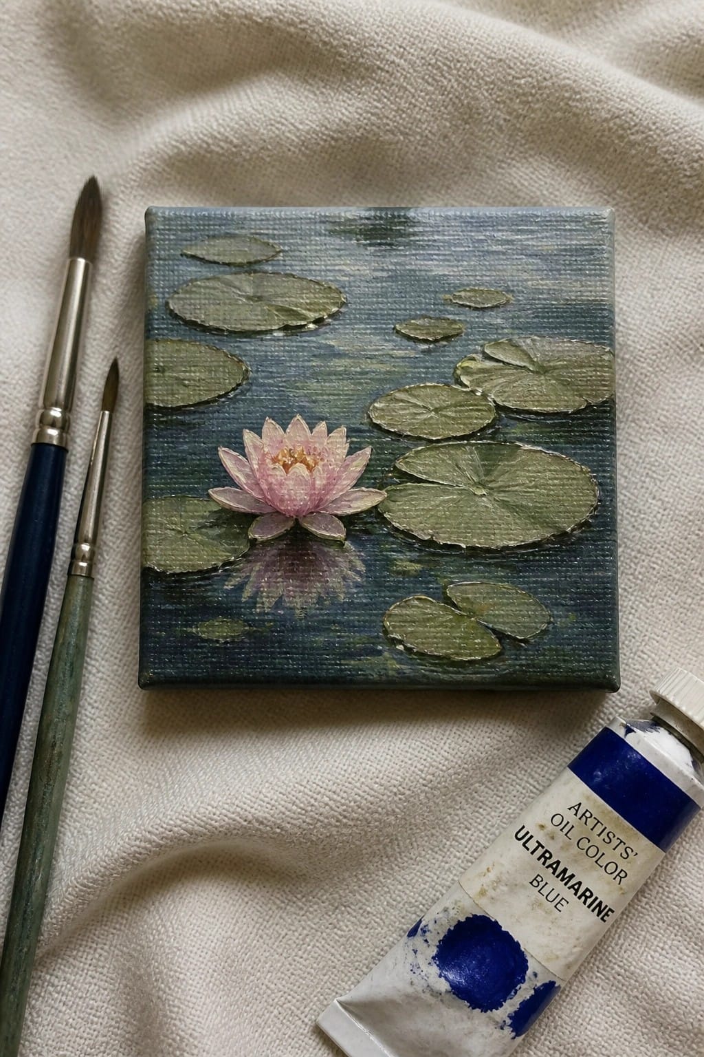 Small canvas with pink water lily, green pads, two brushes, and ultramarine paint tube on fabric.