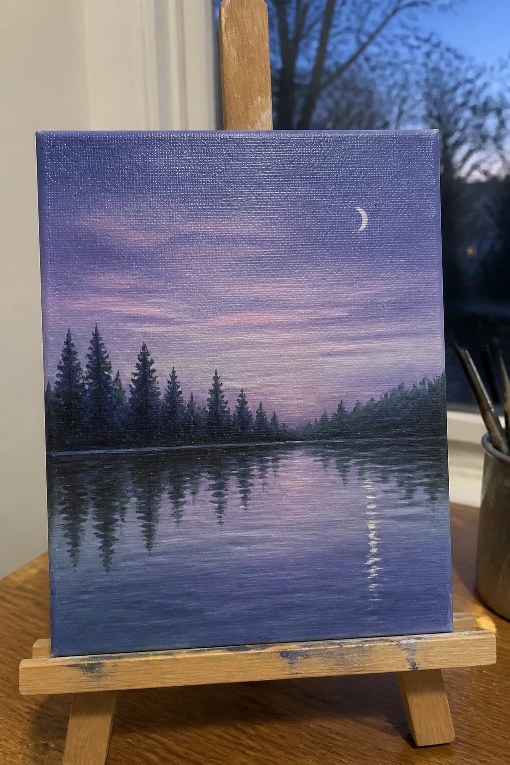 Painting on easel depicts crescent moon over purple sky with reflected pine trees in water.